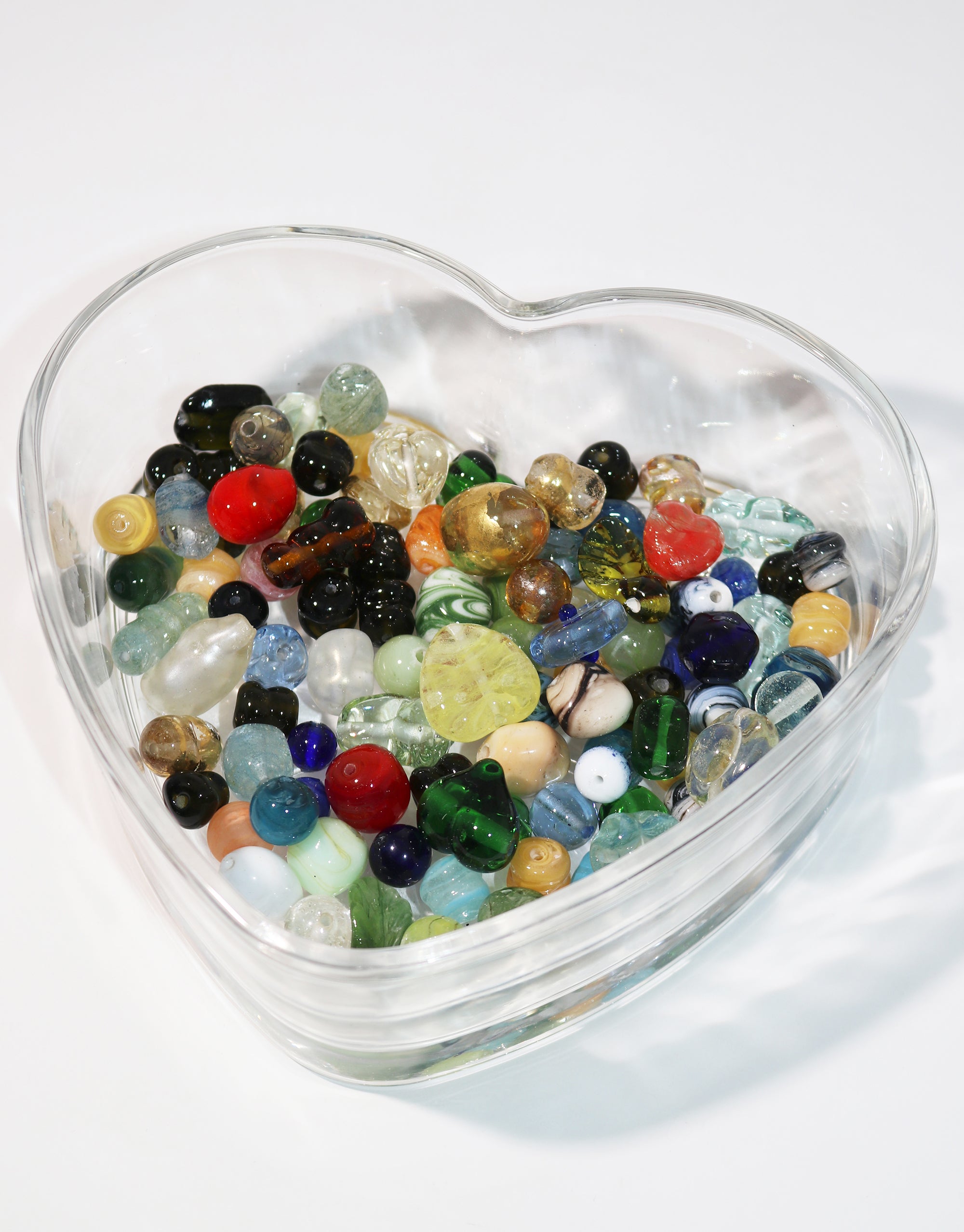 Heart-shaped glass bowl filled with assorted colorful glass marbles on a white surface.