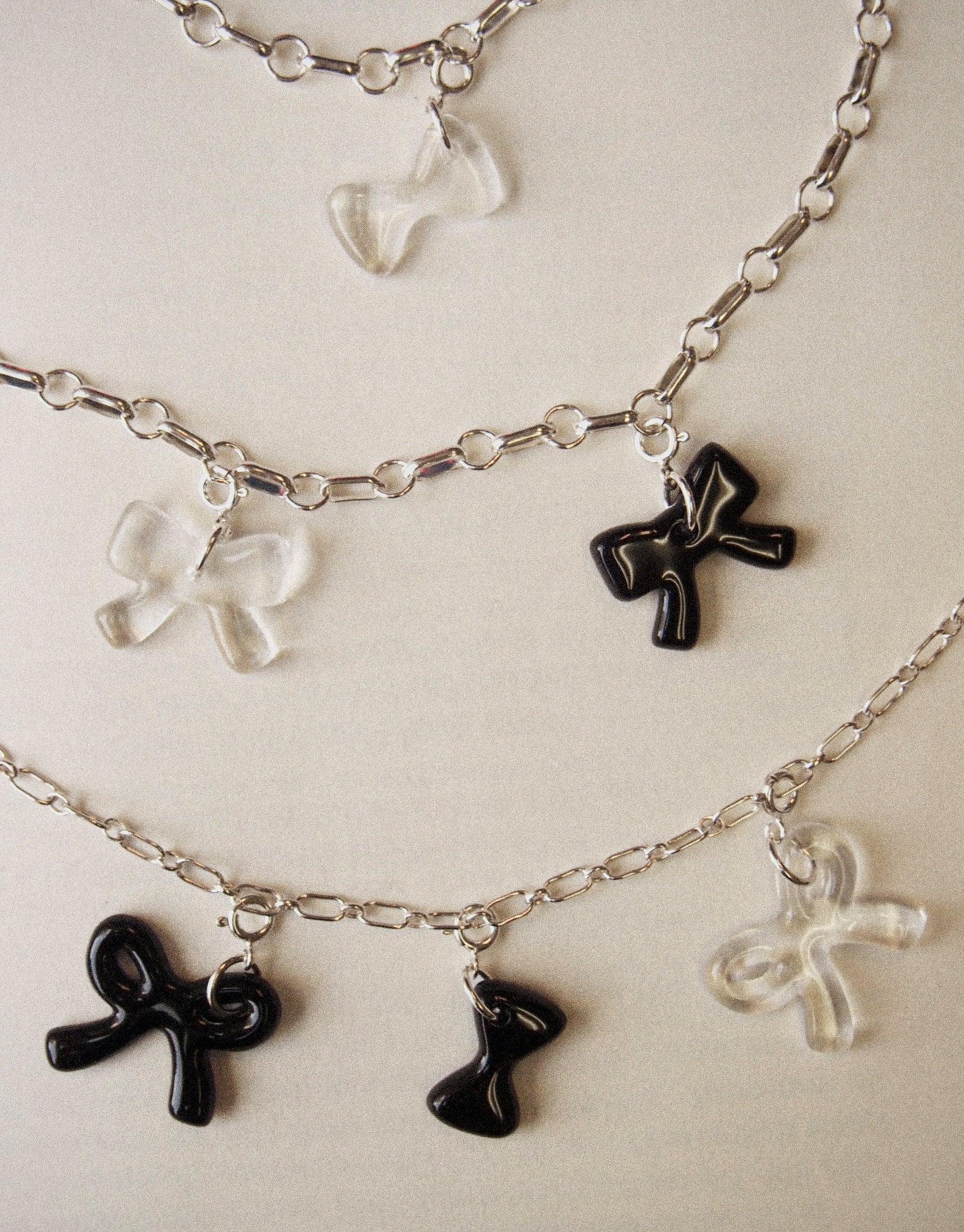 Silver chain necklaces with transparent and black bow-shaped charms, showcasing the playful Icon Charm collection. Versatile accessory for layering or wearing alone. Bow A | Icon Charm by CLED.