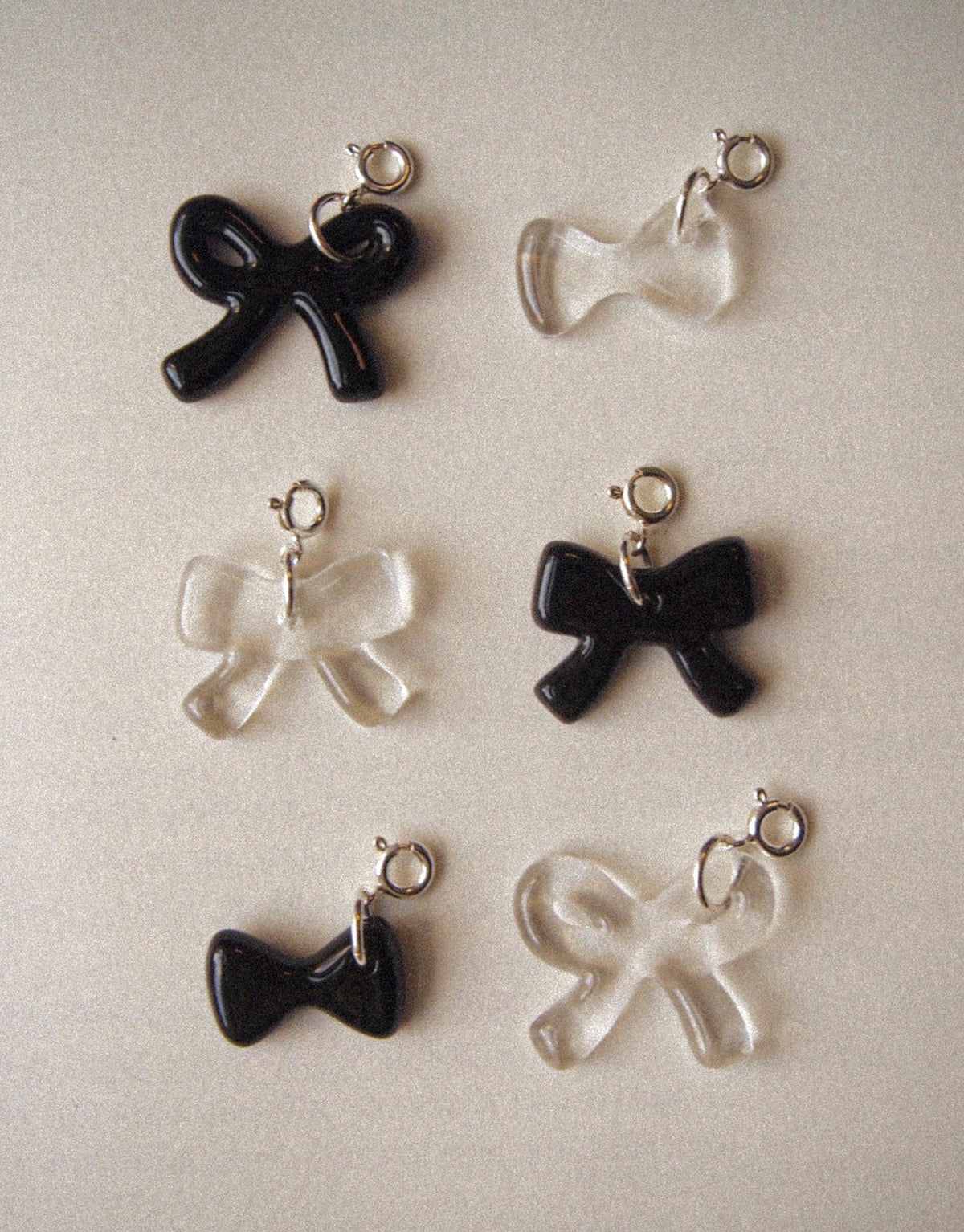 Assortment of black and clear bow-shaped Icon Charms for jewelry customization. Various sizes and styles of bow charms with attached loops for easy addition to necklaces or bracelets. Bow A Icon Charm showcases playful and distinctive designs for personalized accessories. CLED