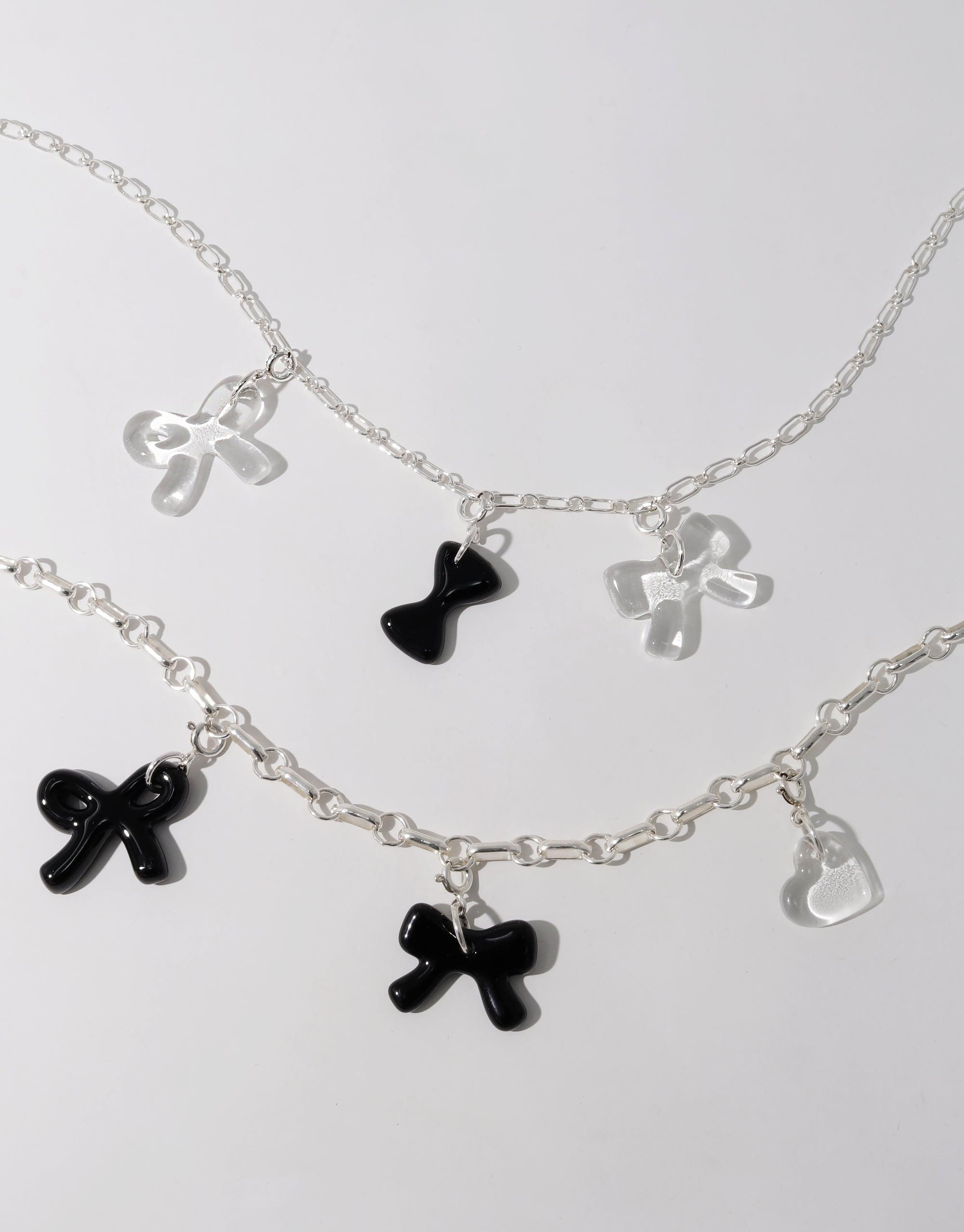 Delicate silver chain necklaces adorned with charming bow-shaped pendants in black and clear, showcasing the playful "Bow A | Icon Charm" design. Perfect for layering or wearing solo, these necklaces add a touch of whimsy to any outfit. CLED