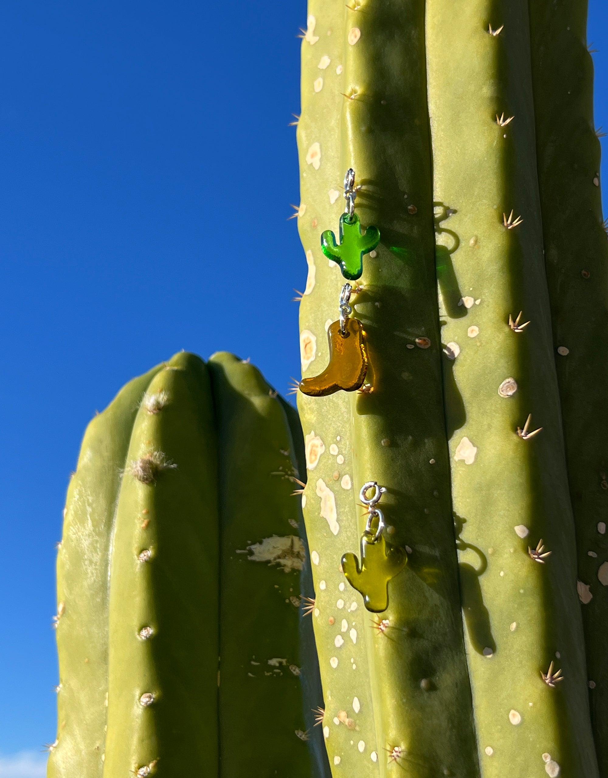 Green cactus icon charm and gold boot charm hanging on large saguaro cactus against vivid blue sky, showcasing Cactus Icon Charm product by CLED