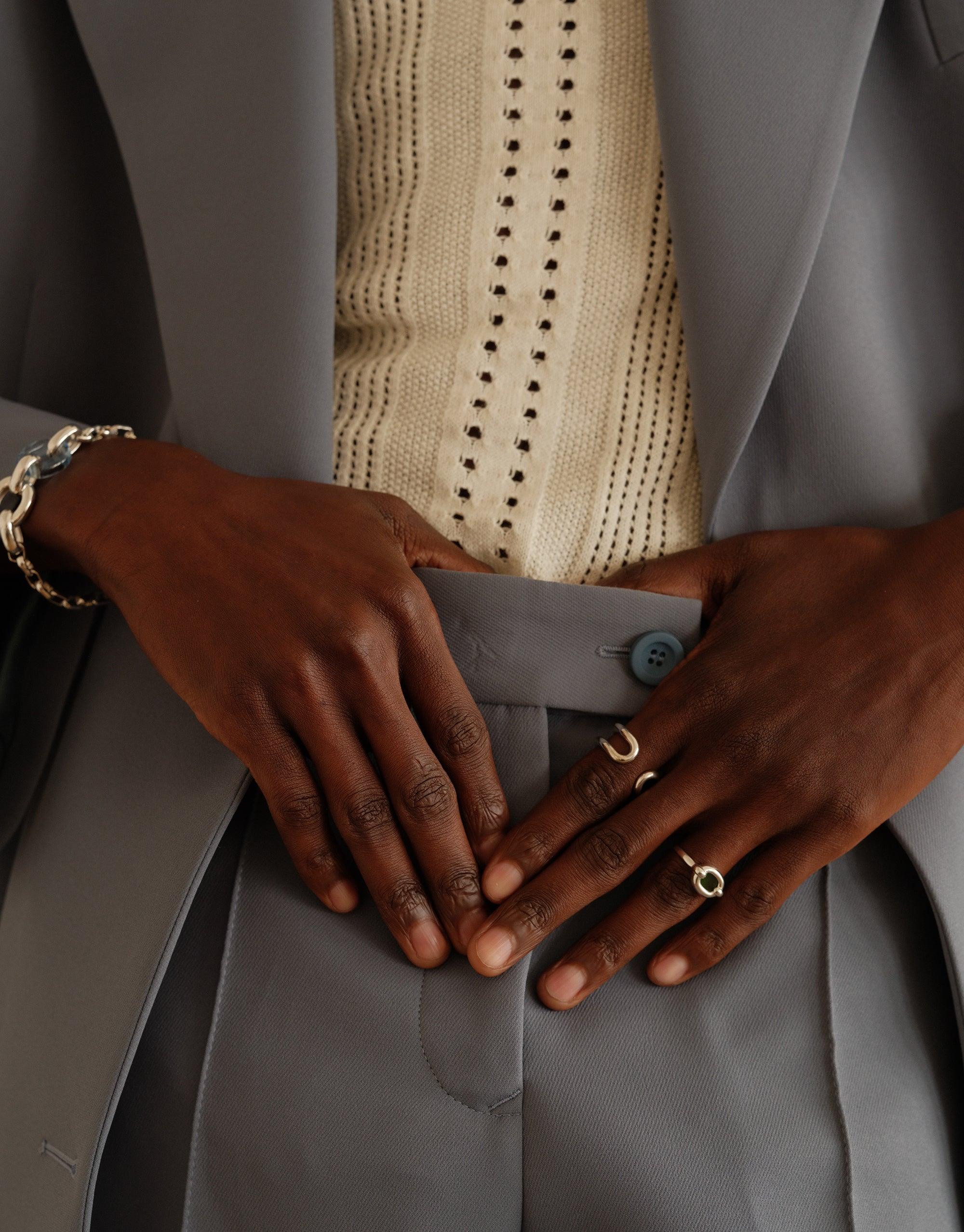 Elegant hands adorned with minimalist jewelry, including a small Core Loop Ring, showcasing sustainable fashion against a backdrop of gray and cream clothing. Upcycled accessories unite style and environmental consciousness. CLED Core Loop Ring | Small