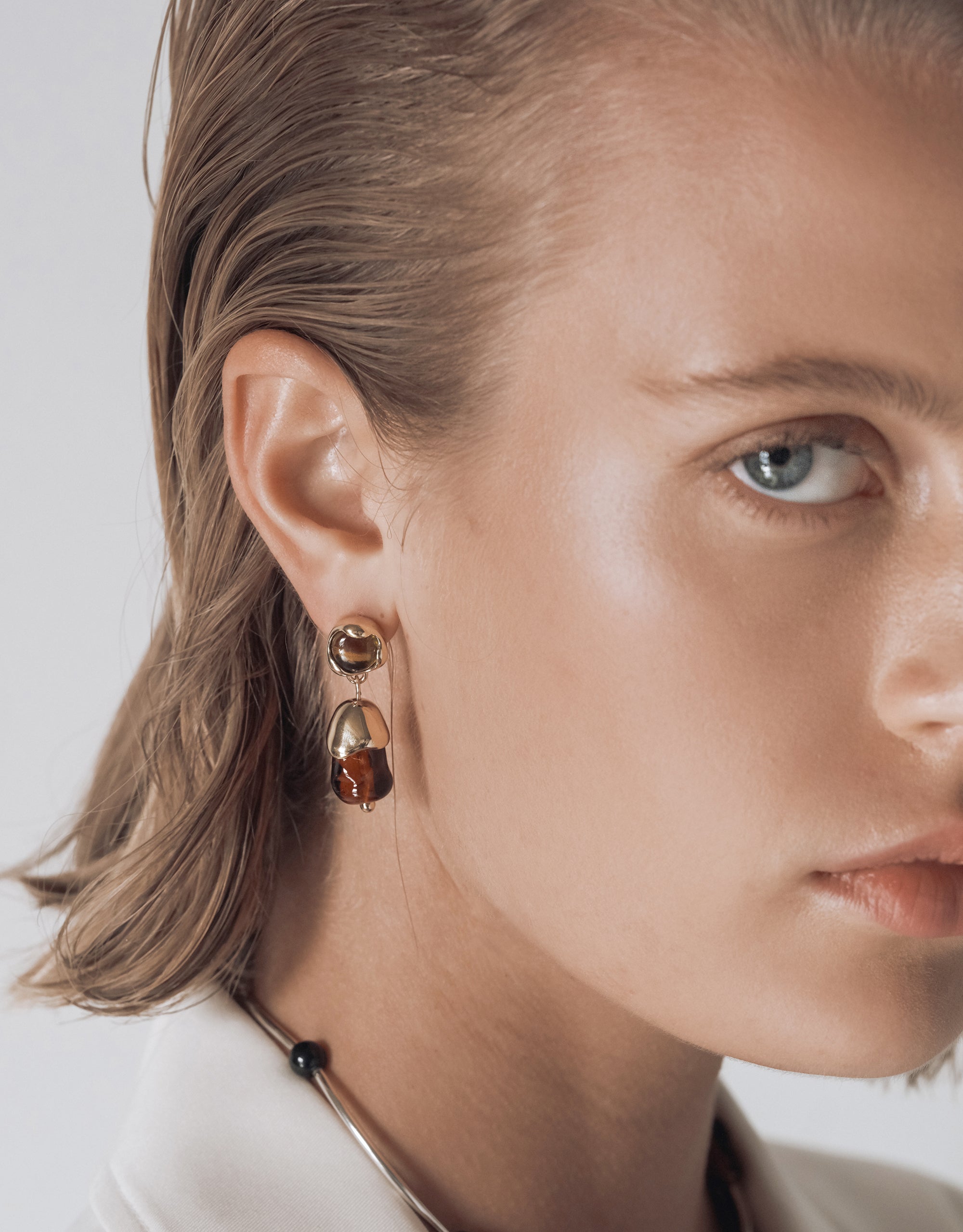 A woman wearing gold and amber-toned glass drop earrings. The earring design includes a smooth gold top element paired with an upcycled glass drop. She also wears a silver choker with a black bead, and her hair is styled slicked back.