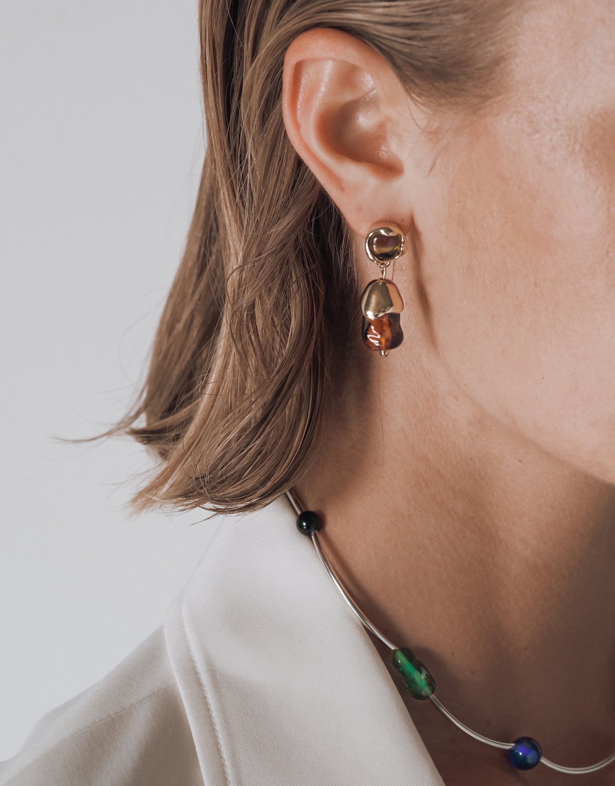 Close-up of a woman's right ear and neck. She is wearing a gold drop earring with two connected pieces—a small round top and a larger amber-toned glass bead below. She also wears a silver choker necklace with a combination of dark and colorful round and organic-shaped upcycled glass beads in black, green, and iridescent blue. Her hair is tucked behind her ear, and she is dressed in a white blazer.