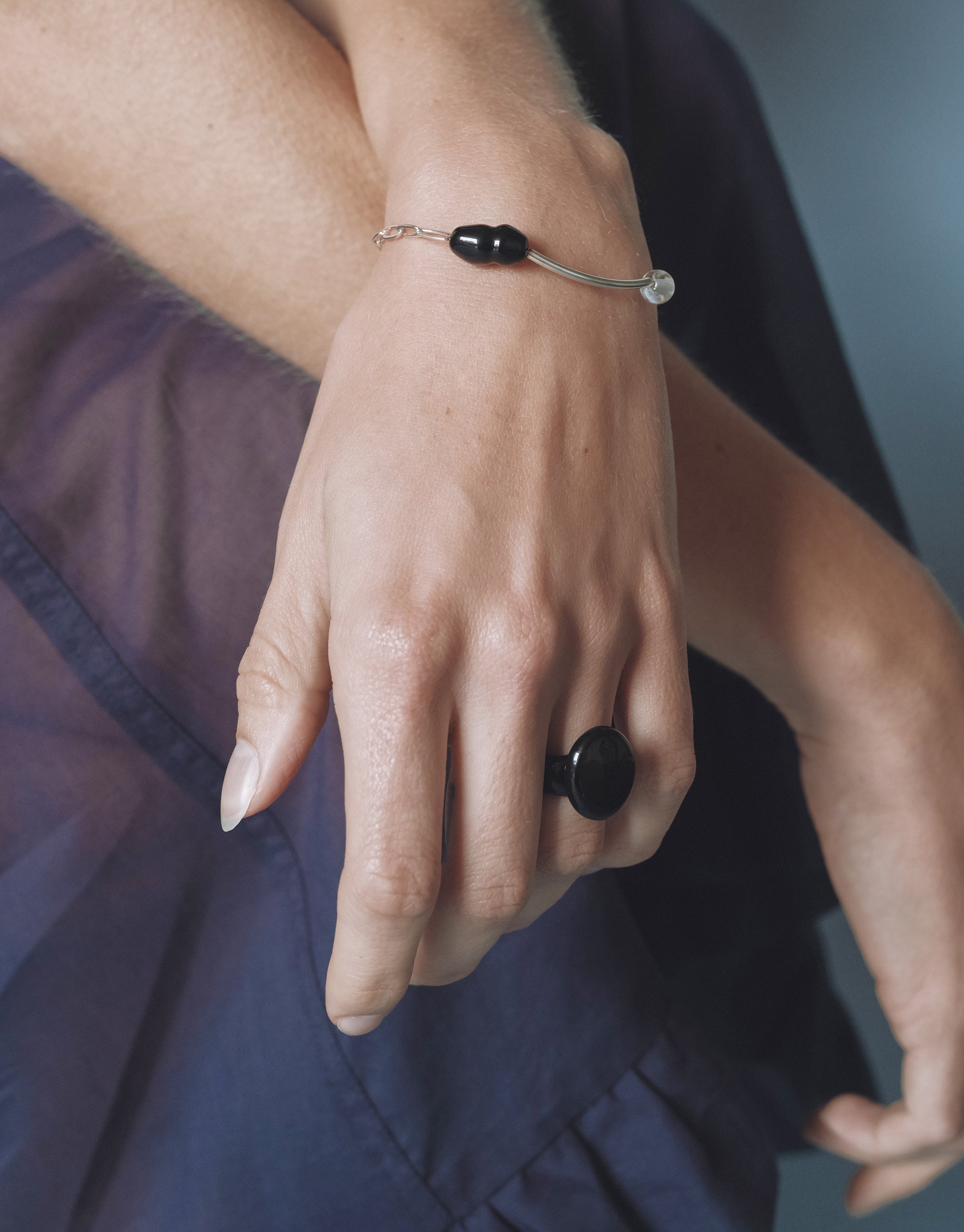 Hand and wrist adorned with a silver bracelet featuring a black Eco Gem and clear bead, paired with a bold black ring, styled against a sheer navy dress.