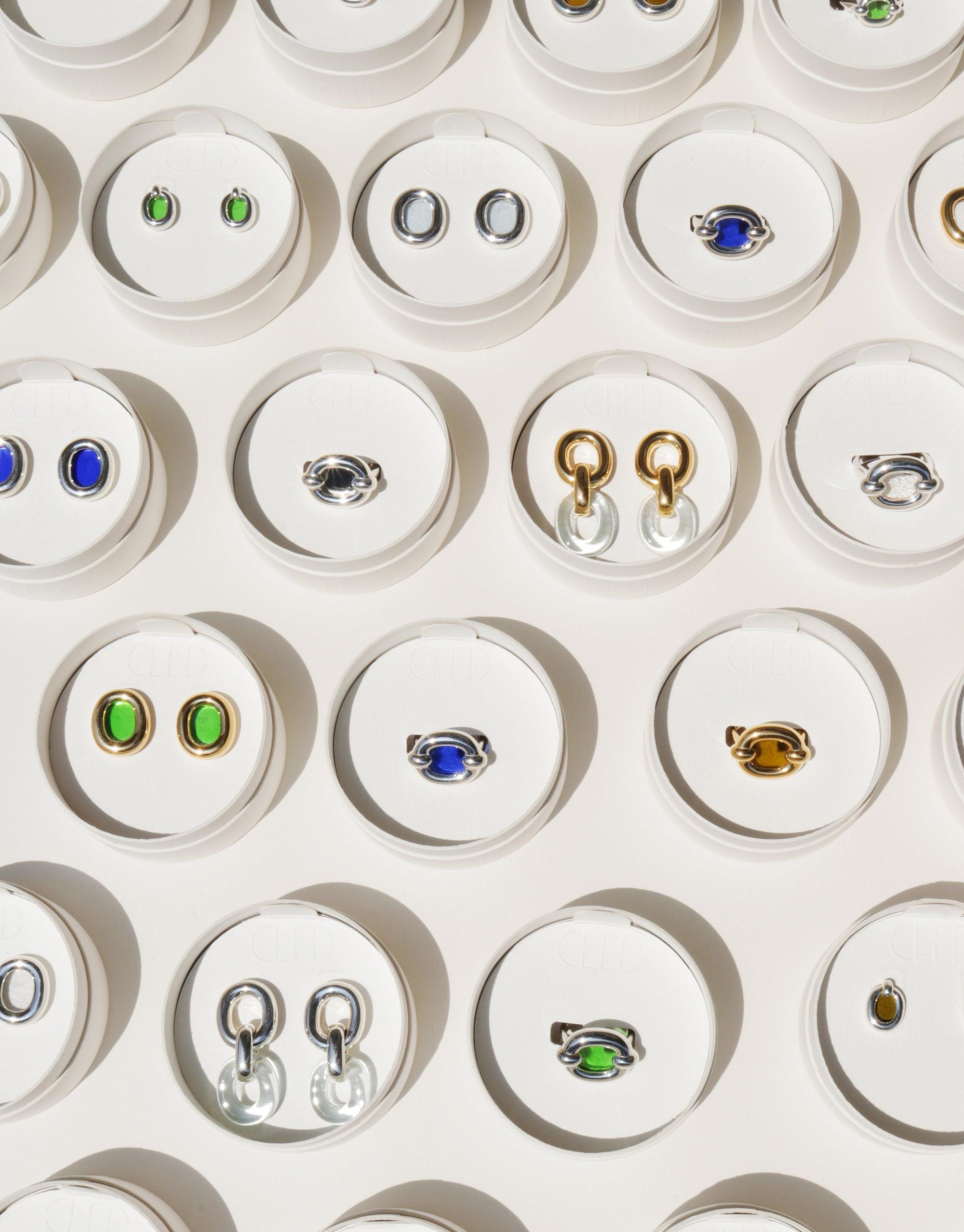 Assorted small earrings displayed in circular white compartments, featuring various designs including loops, studs, and colorful stones. Showcases the diverse styles of the Core Loop Earrings | Small collection, emphasizing upcycled materials and unity. CLED