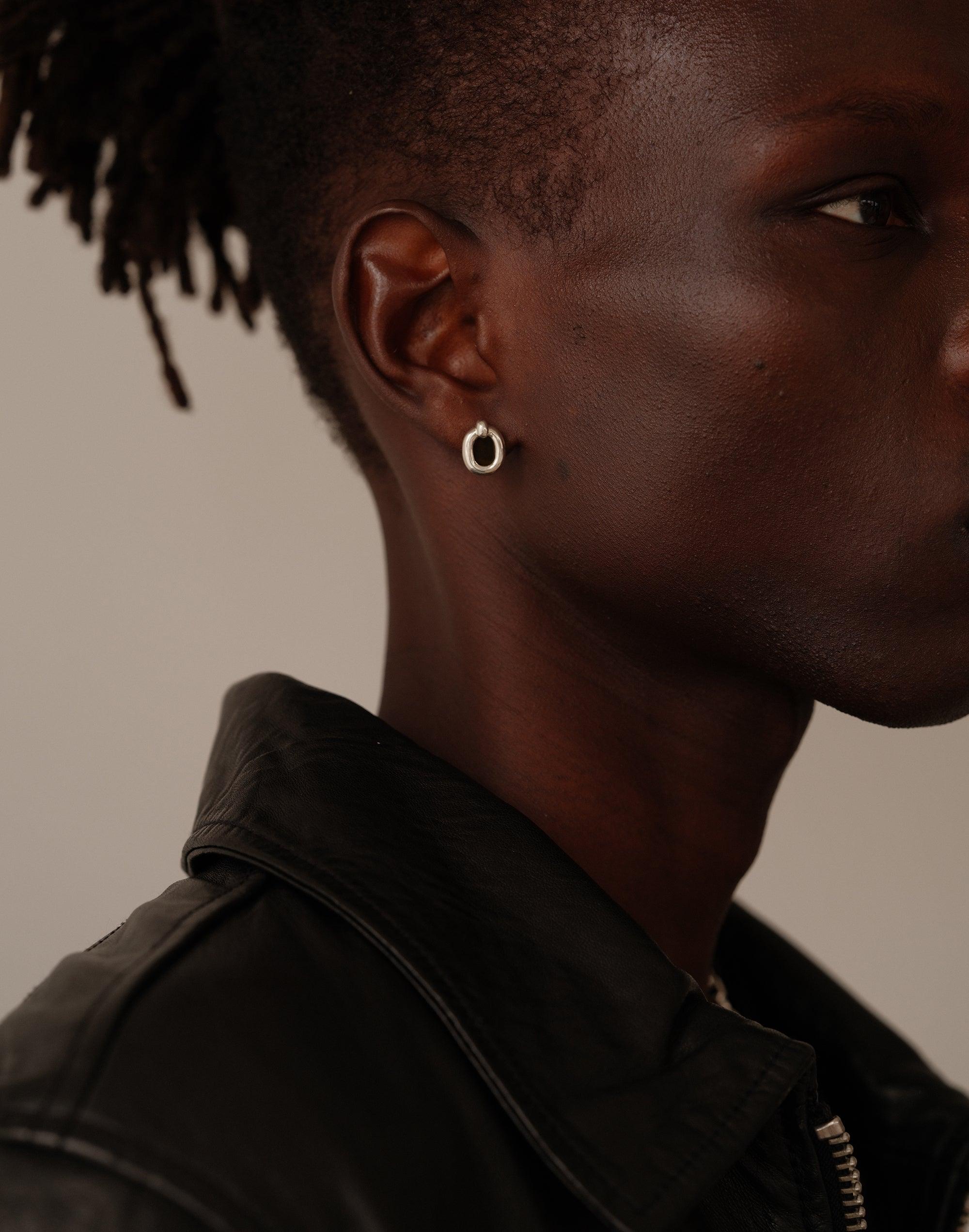 Close-up side profile of model wearing small silver loop earring, showcasing the elegant design against dark skin. The earring complements the model's short hair and black leather jacket, emphasizing its versatility and style. Core Loop Earrings | Small by CLED.