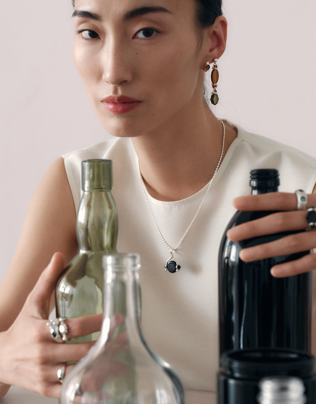 A woman wearing a delicate silver necklace with a dark oval pendant and matching drop earrings, styled with rings, while holding vintage glass bottles.