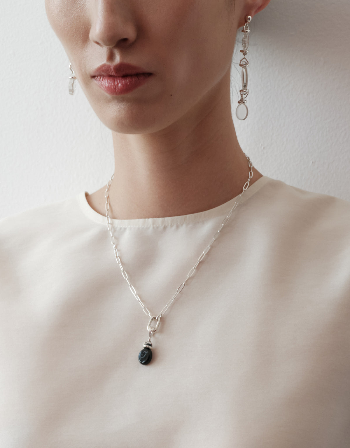 A close-up of a model wearing a silver chain necklace with a dark oval pendant, paired with matching silver drop earrings and a cream-colored top against a neutral light gray background.