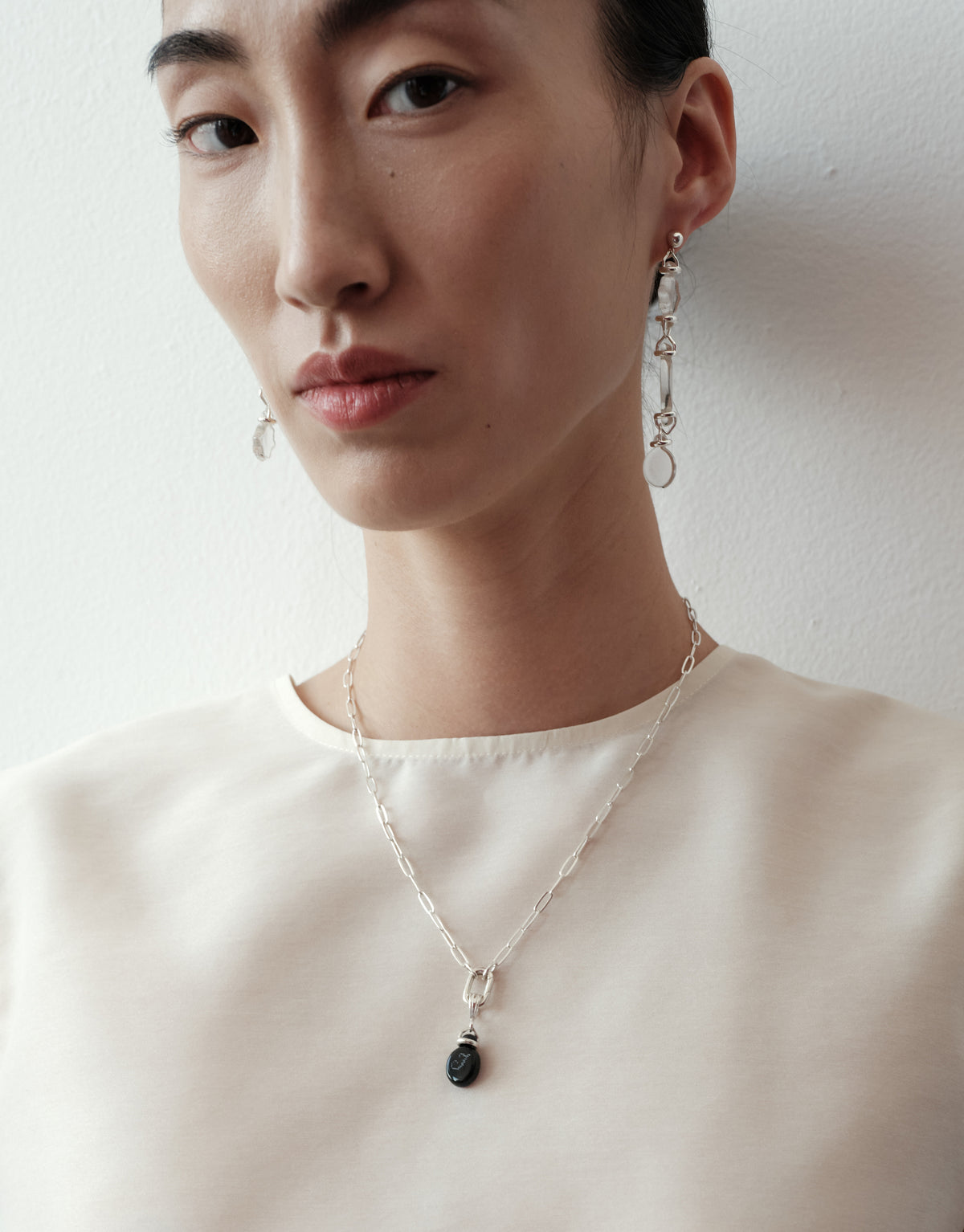 A close-up of a model wearing a delicate silver chain necklace with a dark oval pendant and matching silver drop earrings against a plain white wall.