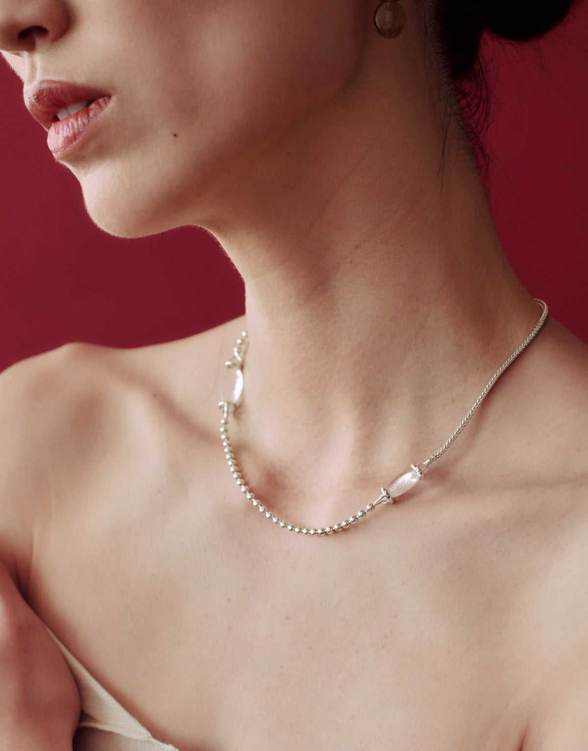 A close-up of a model's neck and upper chest wearing a delicate silver chain necklace with a small clear bead accent, set against a solid dark red background.