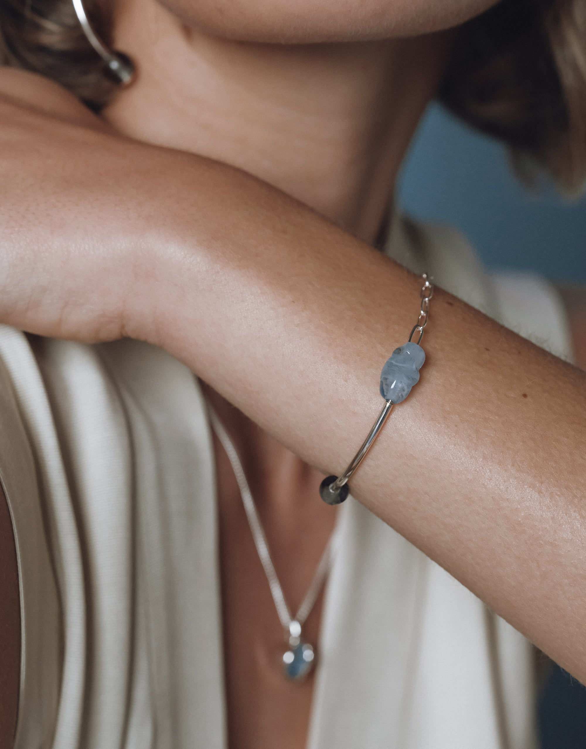 Close-up of a woman resting her chin on her arm, showcasing a silver bracelet with a blue Eco Gem and dark bead, styled with a matching pendant necklace.