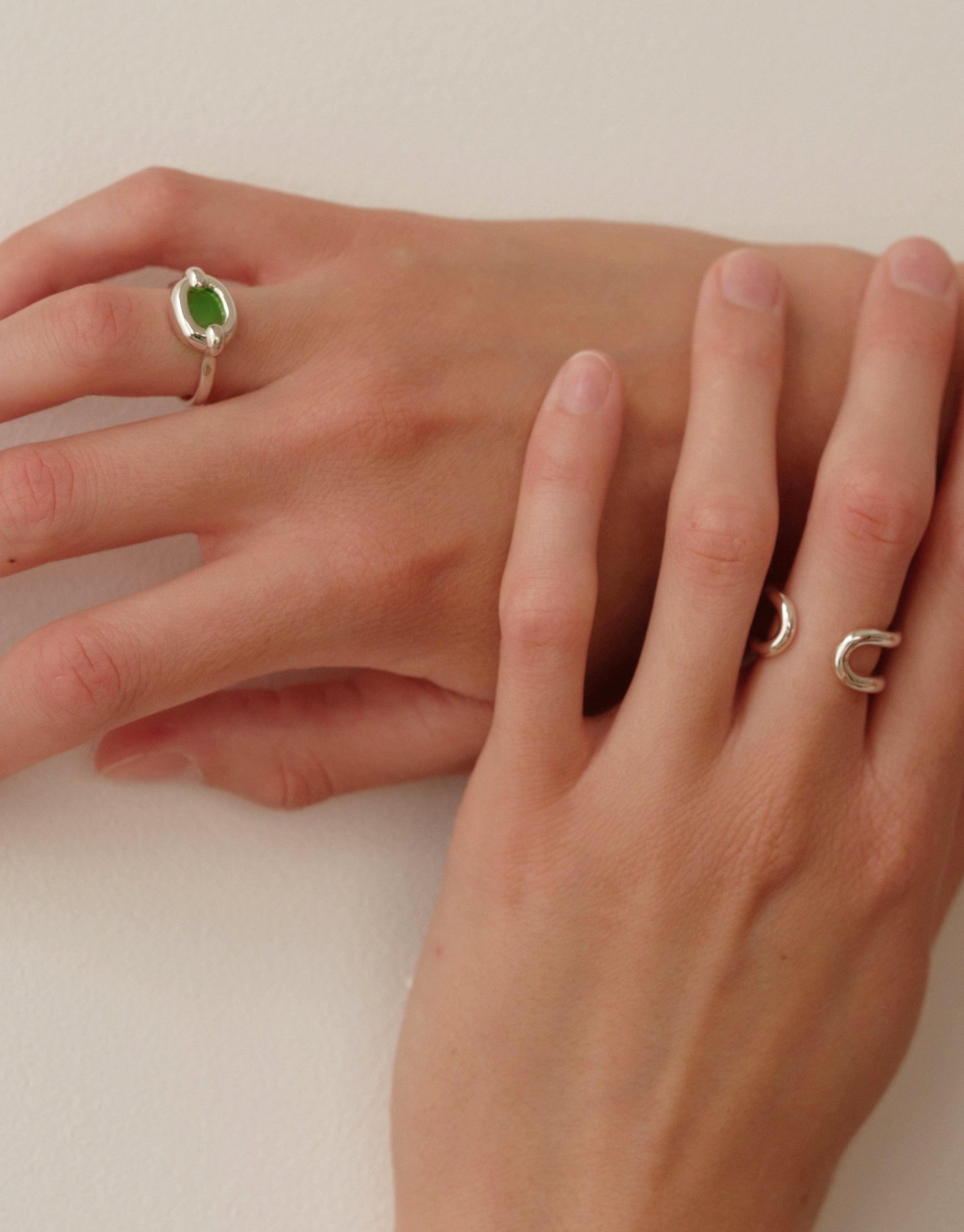 Elegant hands showcasing three unique rings: a silver ring with a green stone and two silver loop rings. The Core Loop Ring | Small is part of CLED's upcycled jewelry collection, symbolizing unity and sustainability. CLED