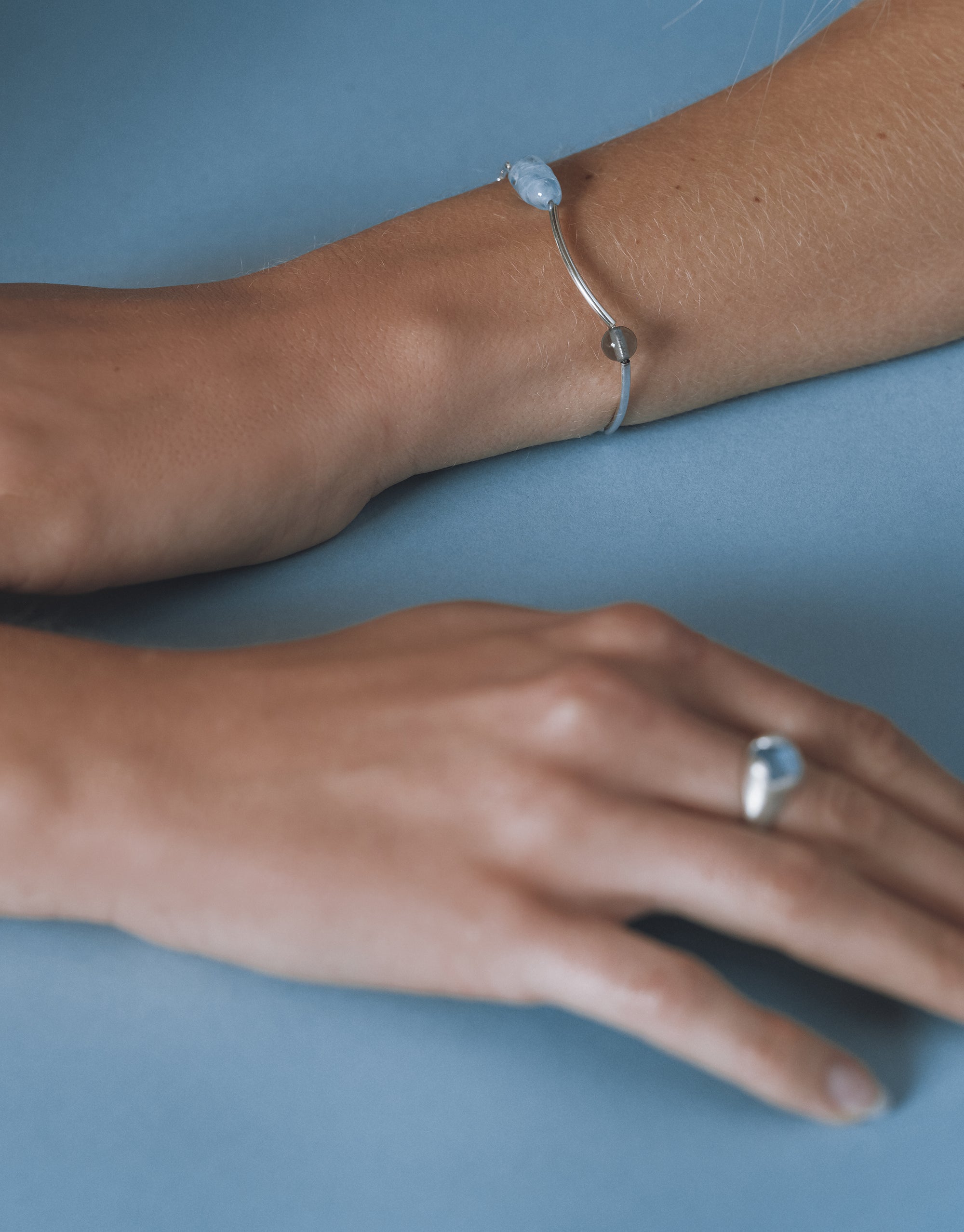 Close-up of a wrist wearing a silver bracelet with a soft blue Eco Gem and a gray bead, styled with a matching silver ring on the hand.