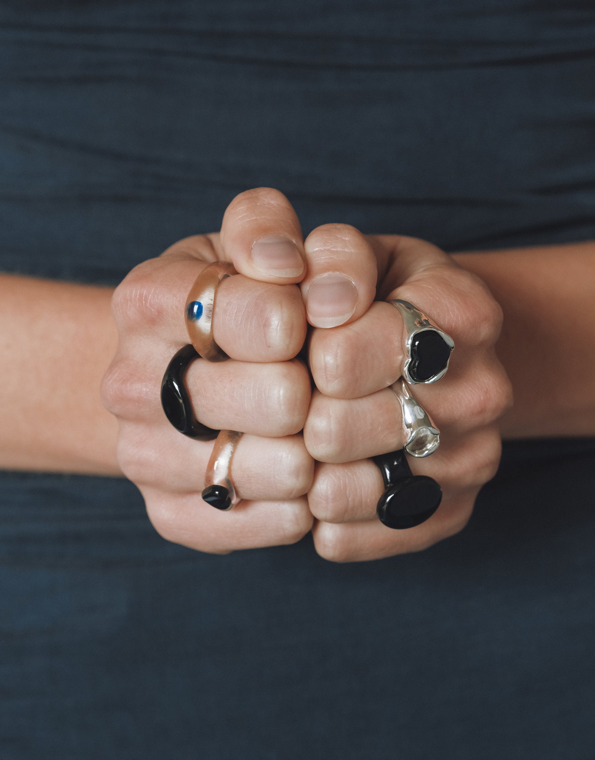 Hands clasped together wearing a variety of bold rings, including black and silver statement rings, a gold band with a blue stone, and mixed textures highlighting individuality and artisanal design.