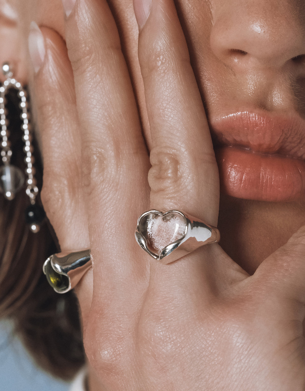 Close up of a model’s hand gently touching her face, showcasing a silver heart shaped ring with a clear Eco Gem and a sculptural silver ring with an olive green stone. A beaded earring with black and clear accents is visible in the background.

