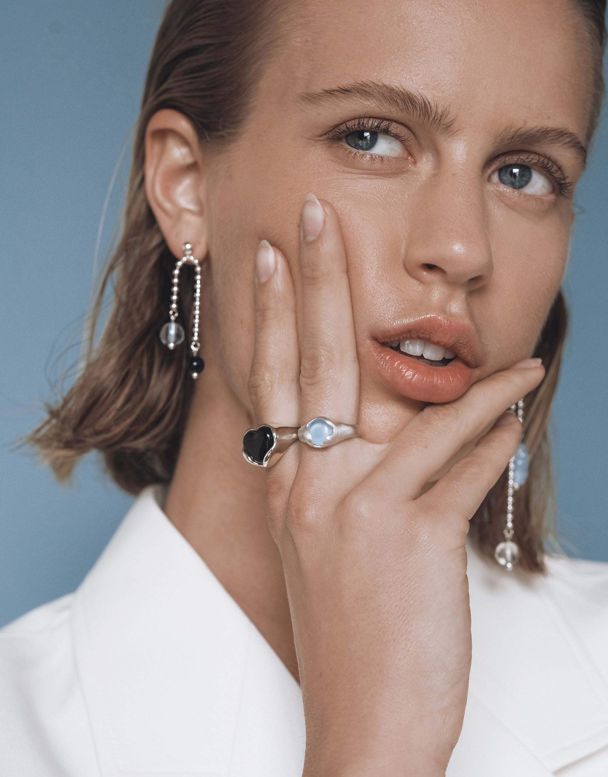 Model wearing Still Earrings featuring asymmetrical beaded chains in silver with matching rings against blue backdrop - CLED