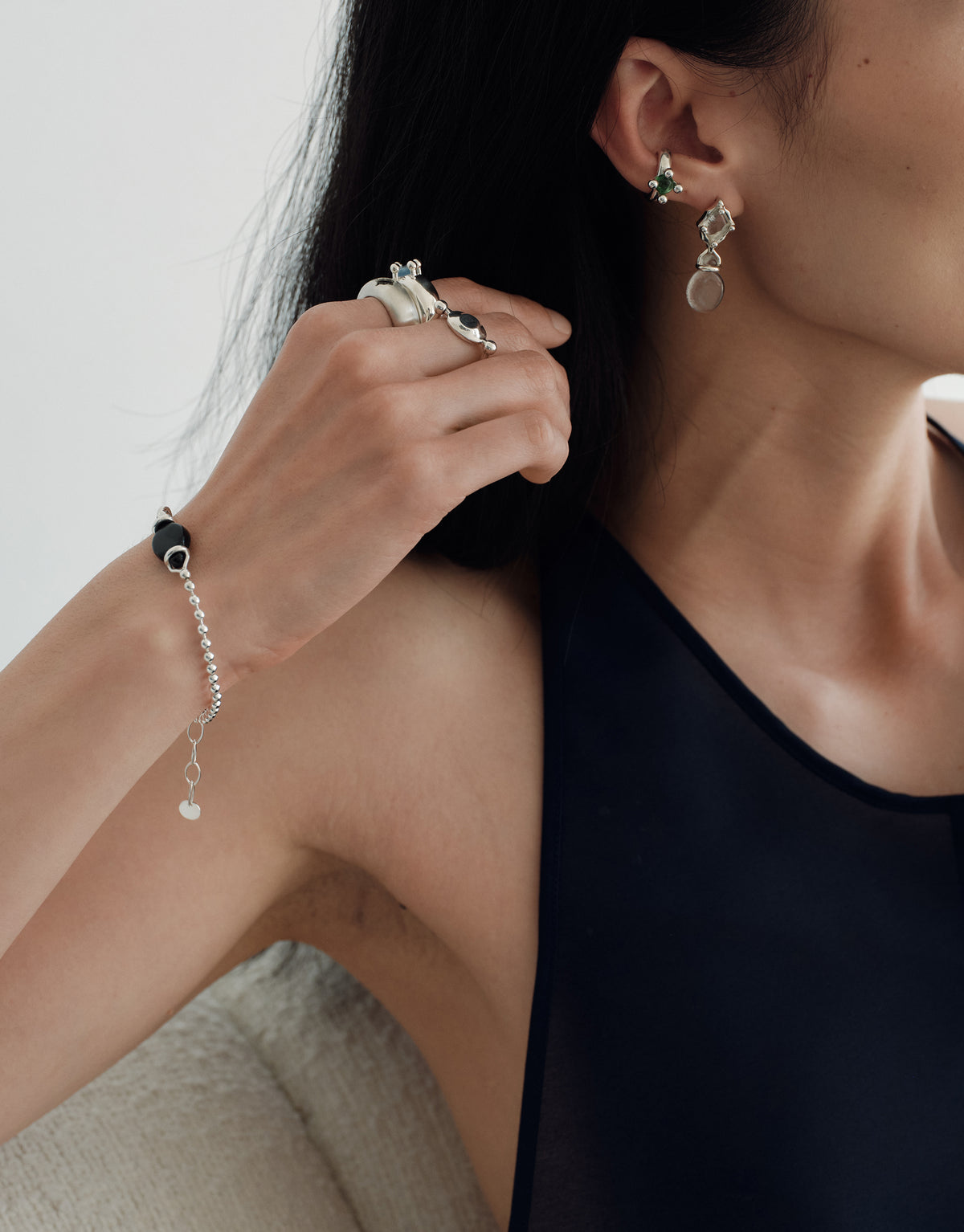 A close-up lifestyle shot of a model wearing a delicate silver beaded chain bracelet with a small heart charm on her wrist, alongside matching silver rings and earrings.