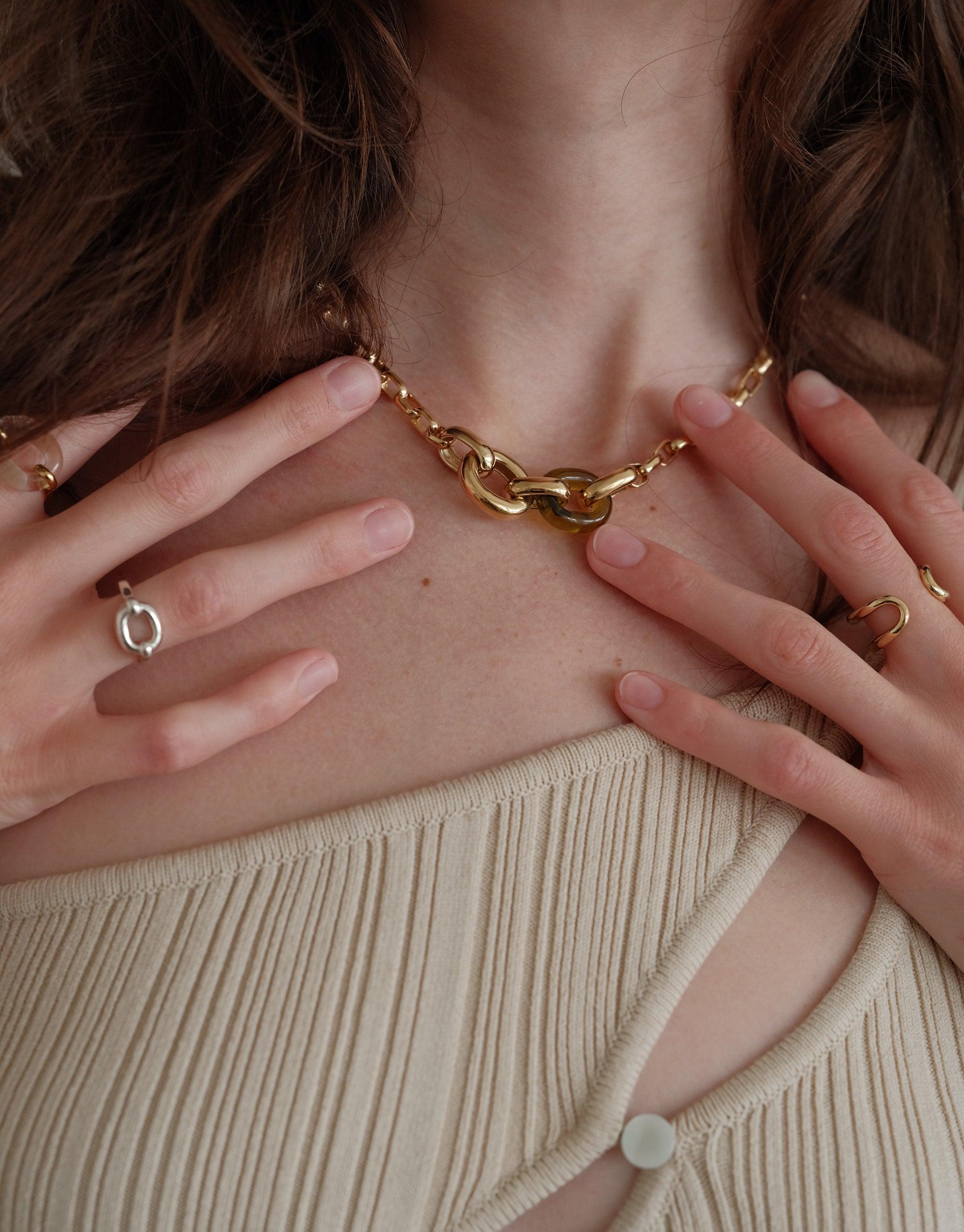 Close-up of a model wearing the Loop Chain Necklace, showcasing its elegant gold links against the skin. Hands delicately touch the necklace, emphasizing its design. The image captures the necklace's stylish appeal and connection to CLED's upcycling mission. Loop Chain Necklace by CLED.