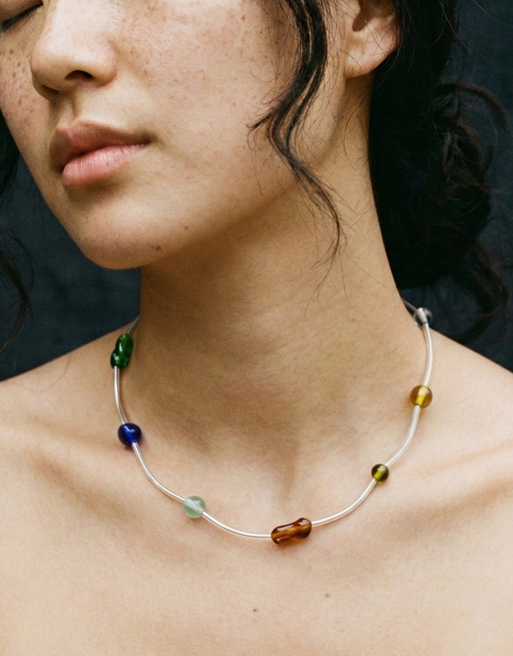 Close-up of a person wearing a delicate necklace with colorful beads against a dark background