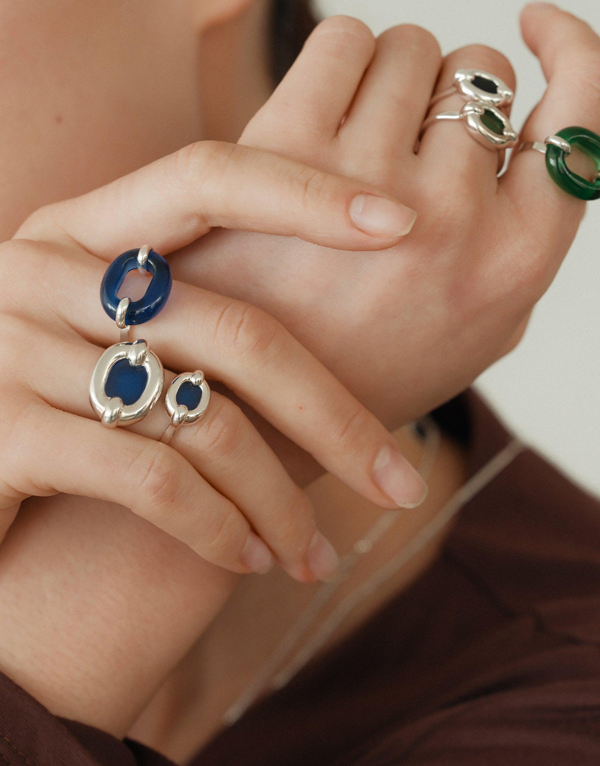 Close-up of hands wearing unique silver rings with blue and green circular elements, showcasing the In The Loop Ring design. Stylish and sustainable jewelry pieces by CLED.