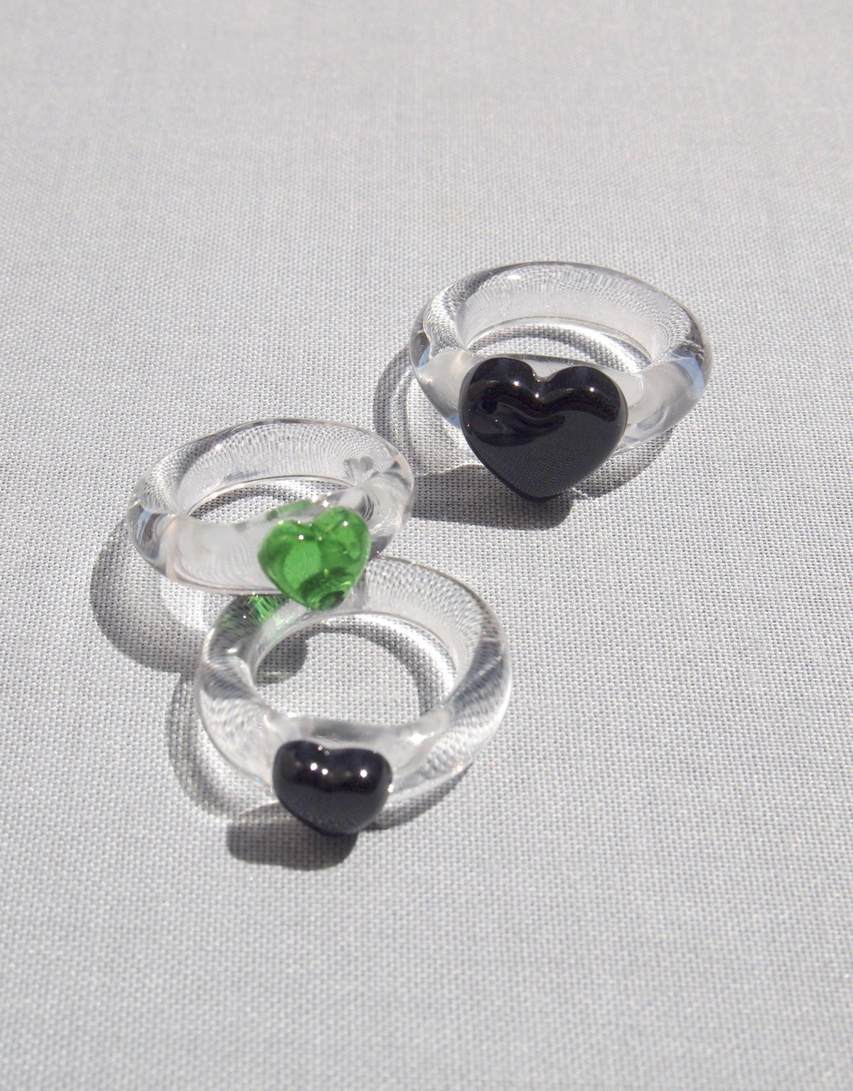 Three transparent glass rings featuring small heart-shaped embellishments in black and green, showcasing the Heart Icon Ring | Small design. Elegant and minimalist jewelry pieces displayed on a light gray background. CLED