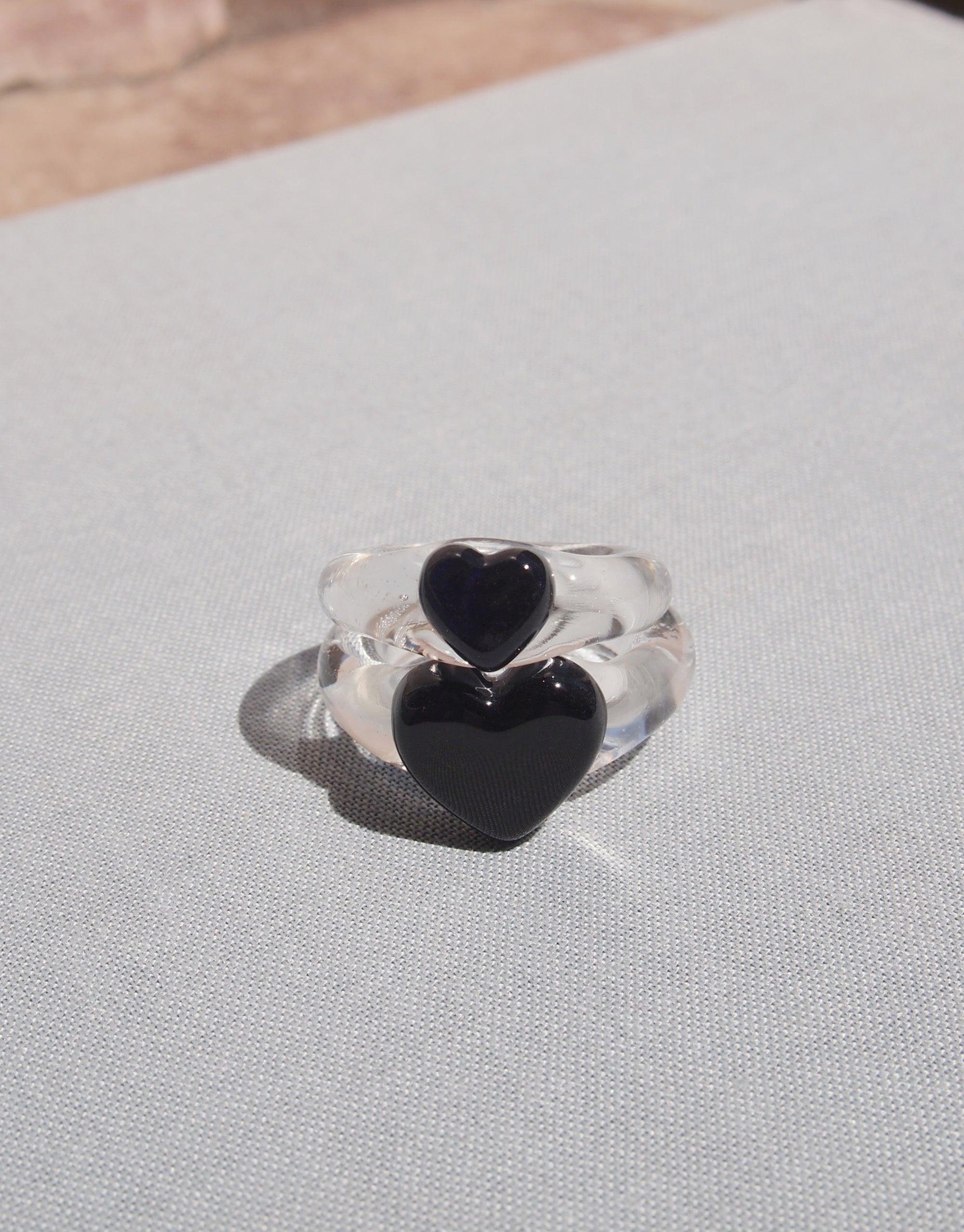 Clear glass ring with two black heart-shaped inlays, one smaller and one larger, resting on a light gray fabric surface. Elegant and minimalist design showcasing the Heart Icon Ring | Small by CLED.