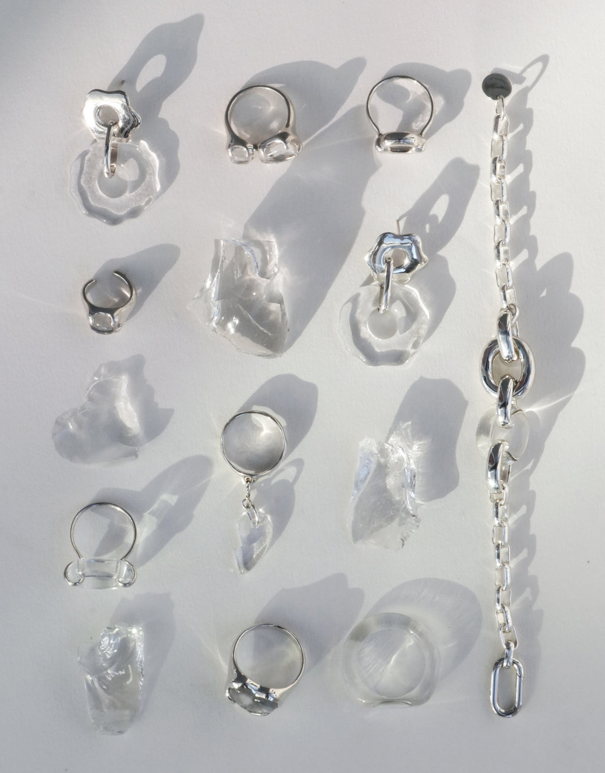 Flat lay of CLED rings, earrings and a chain bracelet in clear upcycled glass and sterling silver, casting soft shadows in daylight