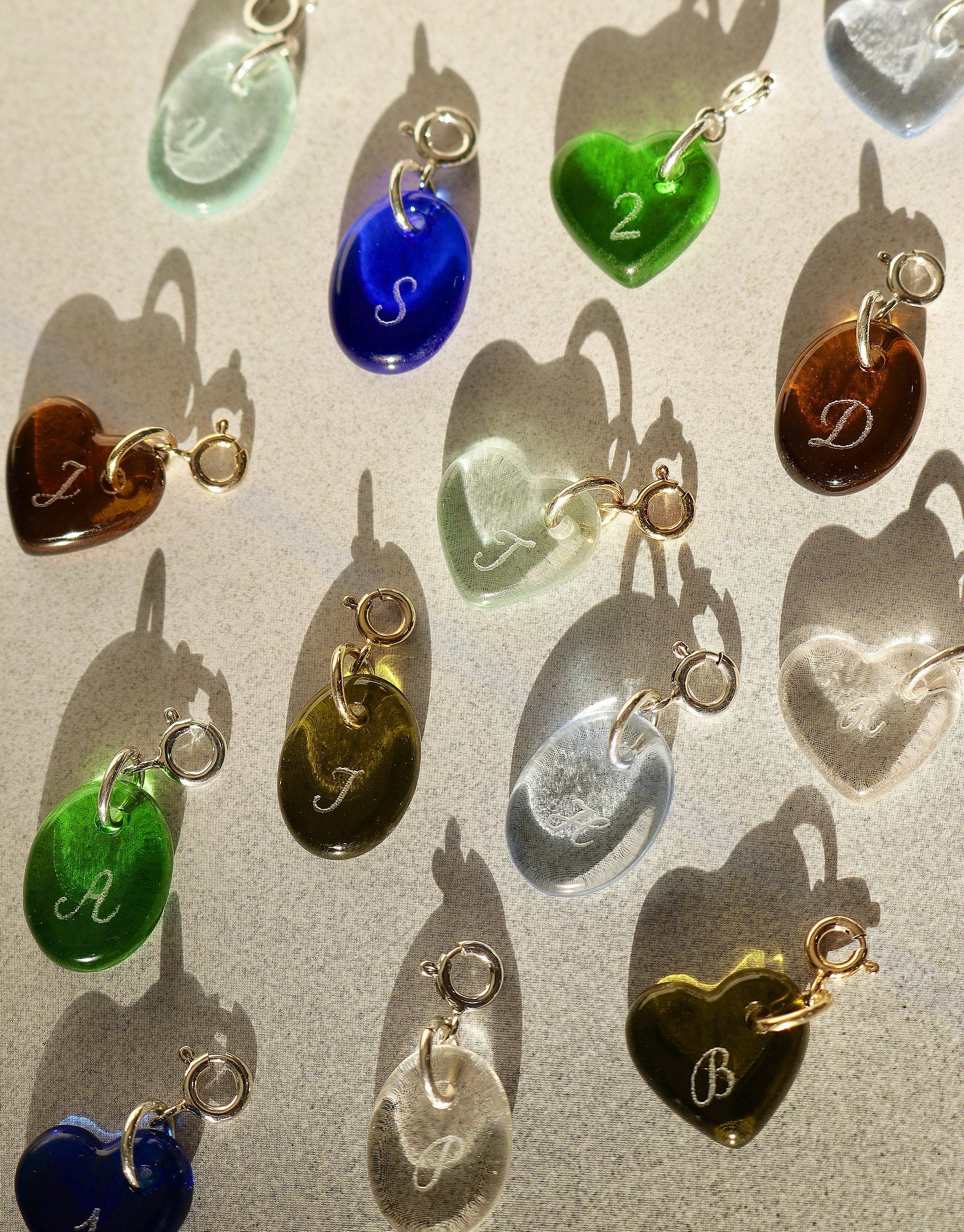 Colorful glass heart and oval-shaped charms with engraved letters, showcasing the Heart | Icon Charm collection. Various hues including blue, green, and amber, arranged on a light background with shadows. Handmade, eco-friendly jewelry pieces by CLED.