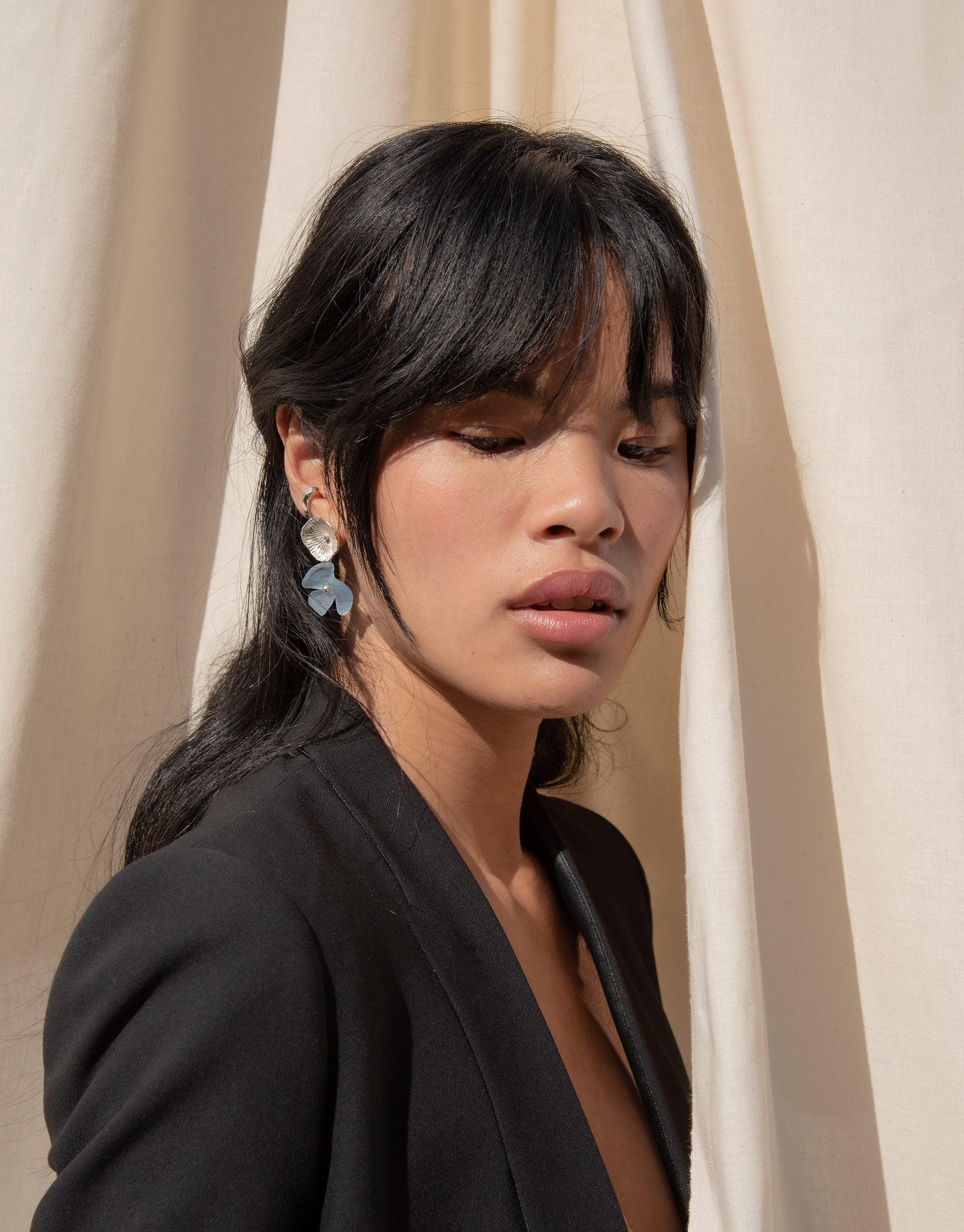 Elegant model wearing Periwinkle Single Earrings, showcasing delicate flower studs with glass petal drops. Earthy and eco-friendly design against a neutral background. Handcrafted sustainable jewelry by CLED.