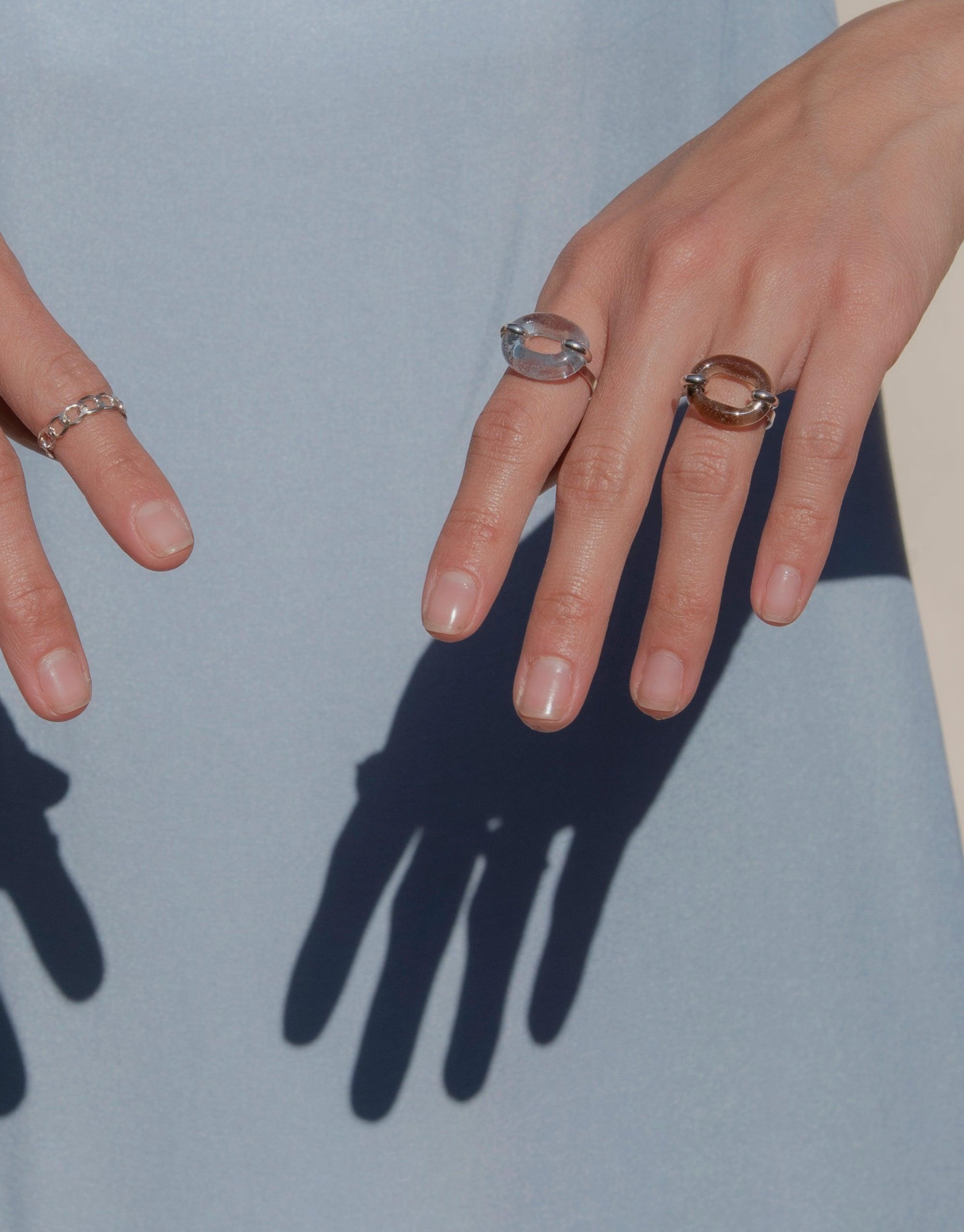 Hands wearing stylish In The Loop Ring, showcasing unique circular design on multiple fingers. Sustainable jewelry symbolizing connection and solidarity. Elegant accessory against light background with shadow detail. CLED