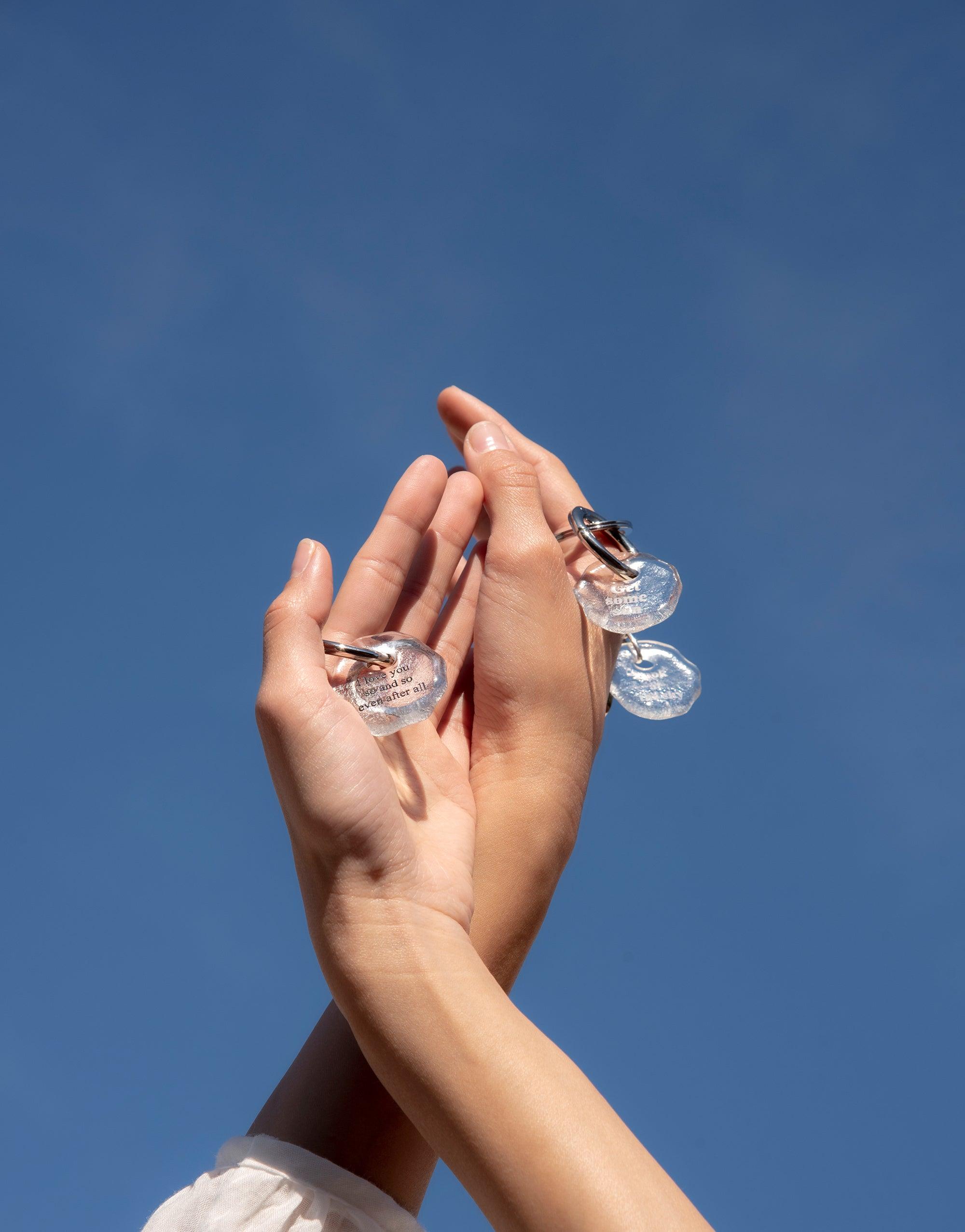 Hands holding up Messenger Keyring | Inhale. Exhale against blue sky. Clear glass charms with engraved messages attached to metal rings. Eco-friendly, handcrafted keyrings showcasing the Messenger Collection's thoughtful design. CLED
