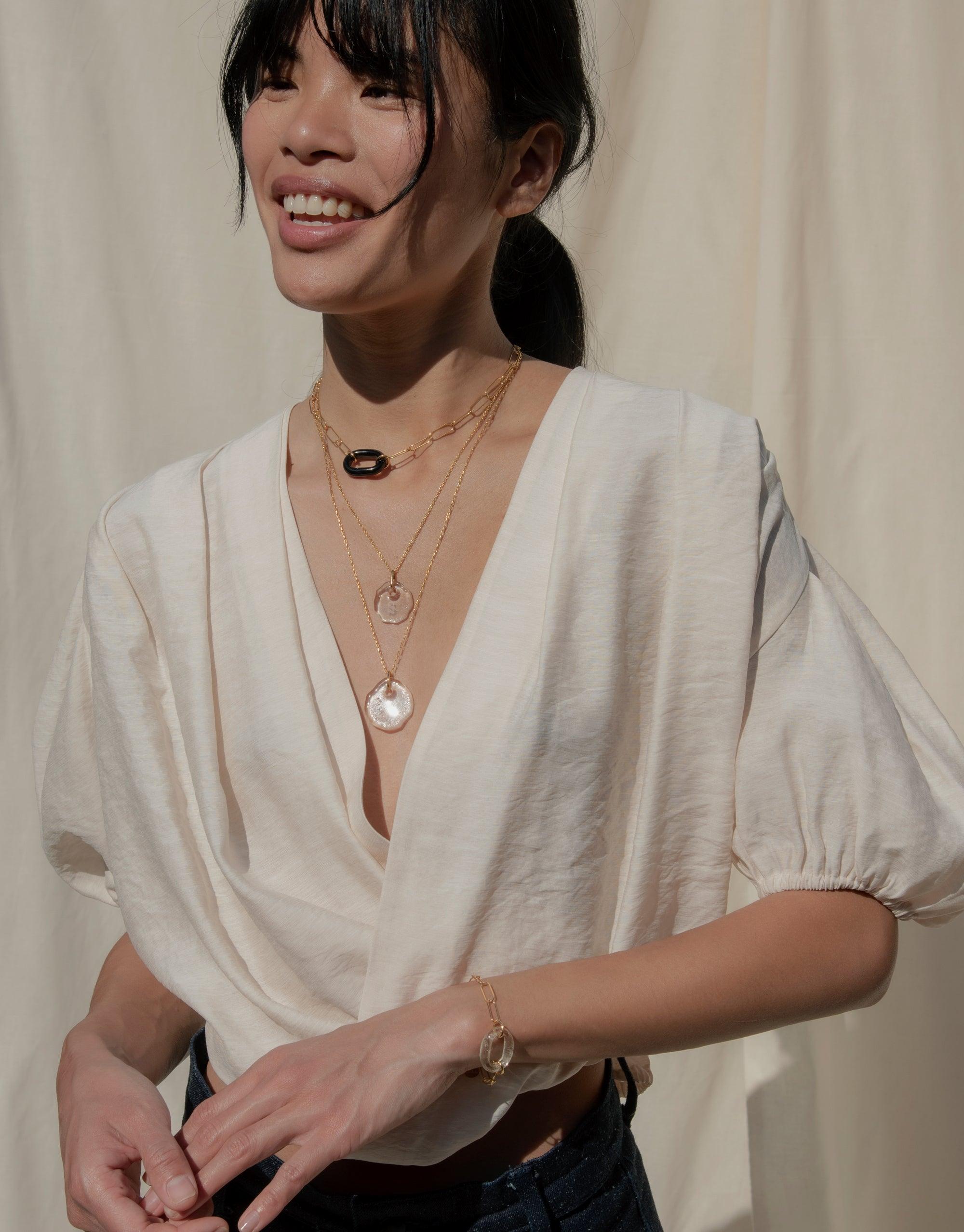 Woman wearing white blouse and layered gold necklaces featuring unique pendants, showcasing the Messenger Necklace with Clear Engraving. Elegant and personalized jewelry for stylish communication. CLED