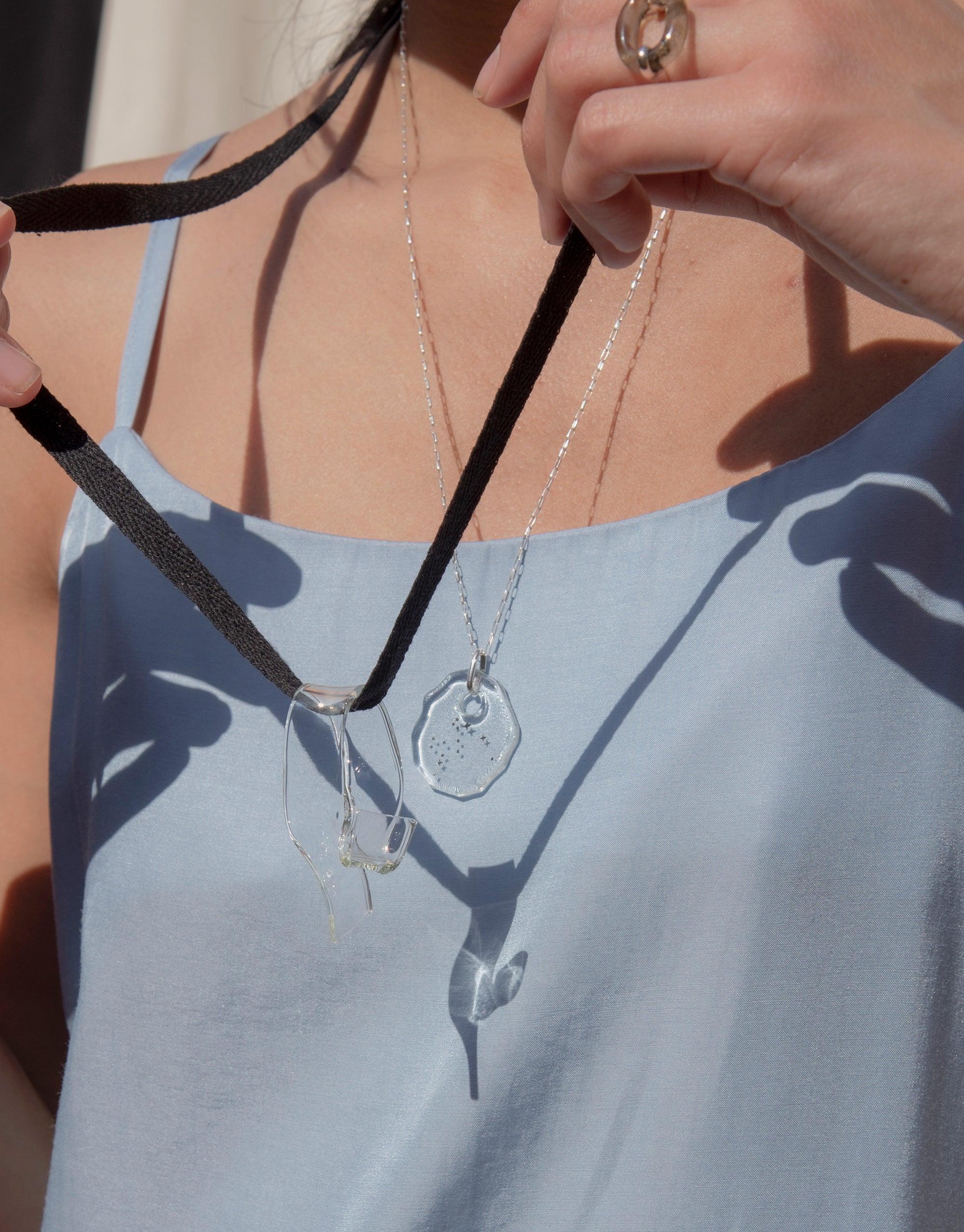 Eco-friendly Ribbon Necklace featuring hand-formed glass pendant on cotton ribbon, showcasing sustainable jewelry design. Close-up of necklace worn with light blue top, highlighting unique serpentine shape and eco-conscious craftsmanship. Ribbon Necklace by CLED.