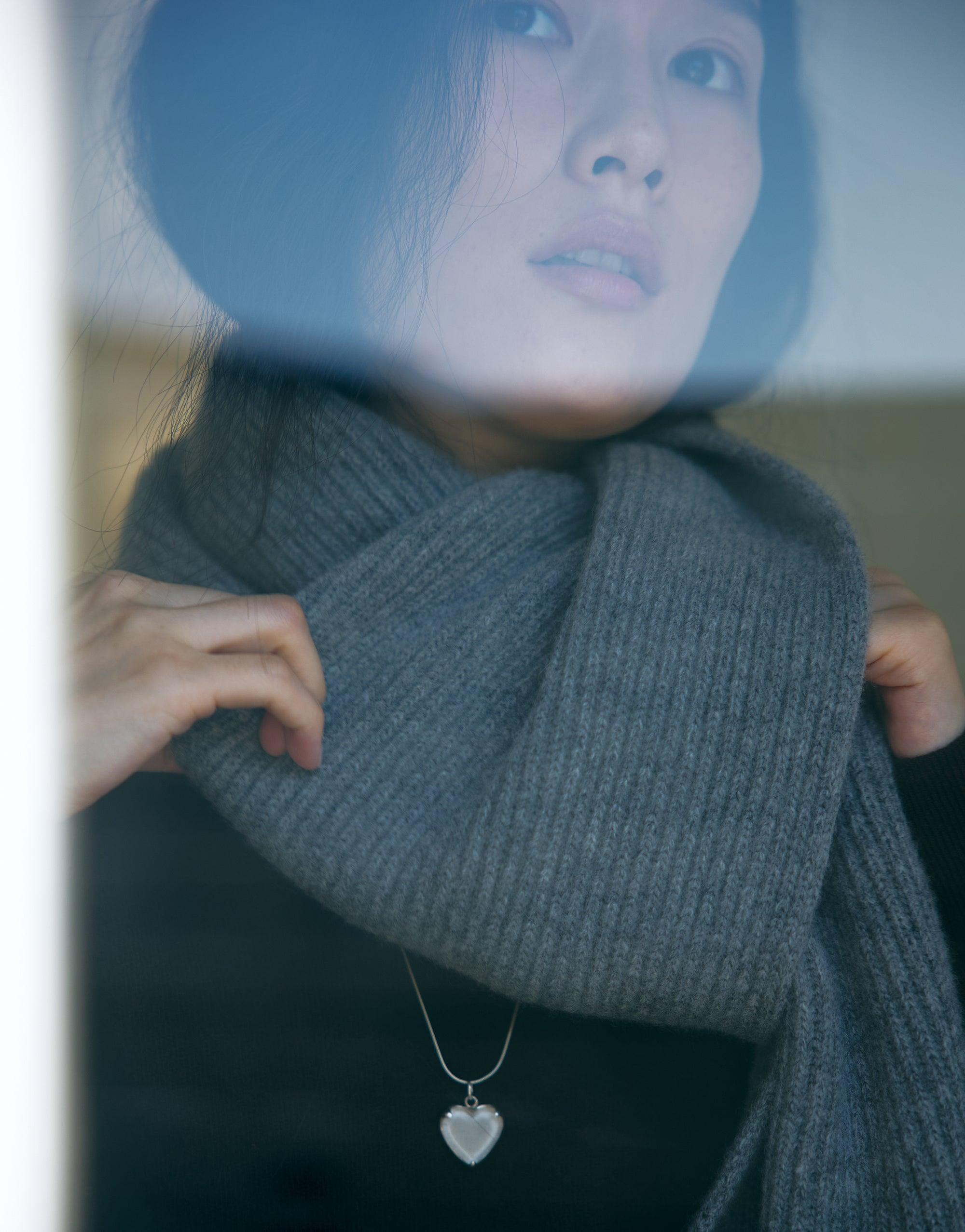 Close-up view of a person wearing a gray knit scarf and the Heart Frame Long Necklace. The delicate heart-shaped pendant is visible against a dark top, showcasing the elegant design of this handmade sterling silver or gold vermeil necklace by CLED.