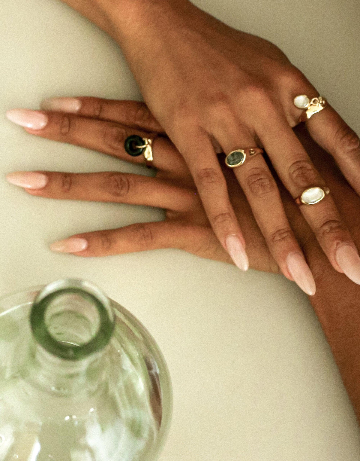 Elegant hand displaying multiple rings, including the Oval Dangling Ring, showcasing eco-friendly charm designs in gold and silver tones. Upcycled glass bottle visible, emphasizing sustainable jewelry craftsmanship. Handmade luxury accessories for conscious fashion enthusiasts. CLED Oval Dangling Ring.