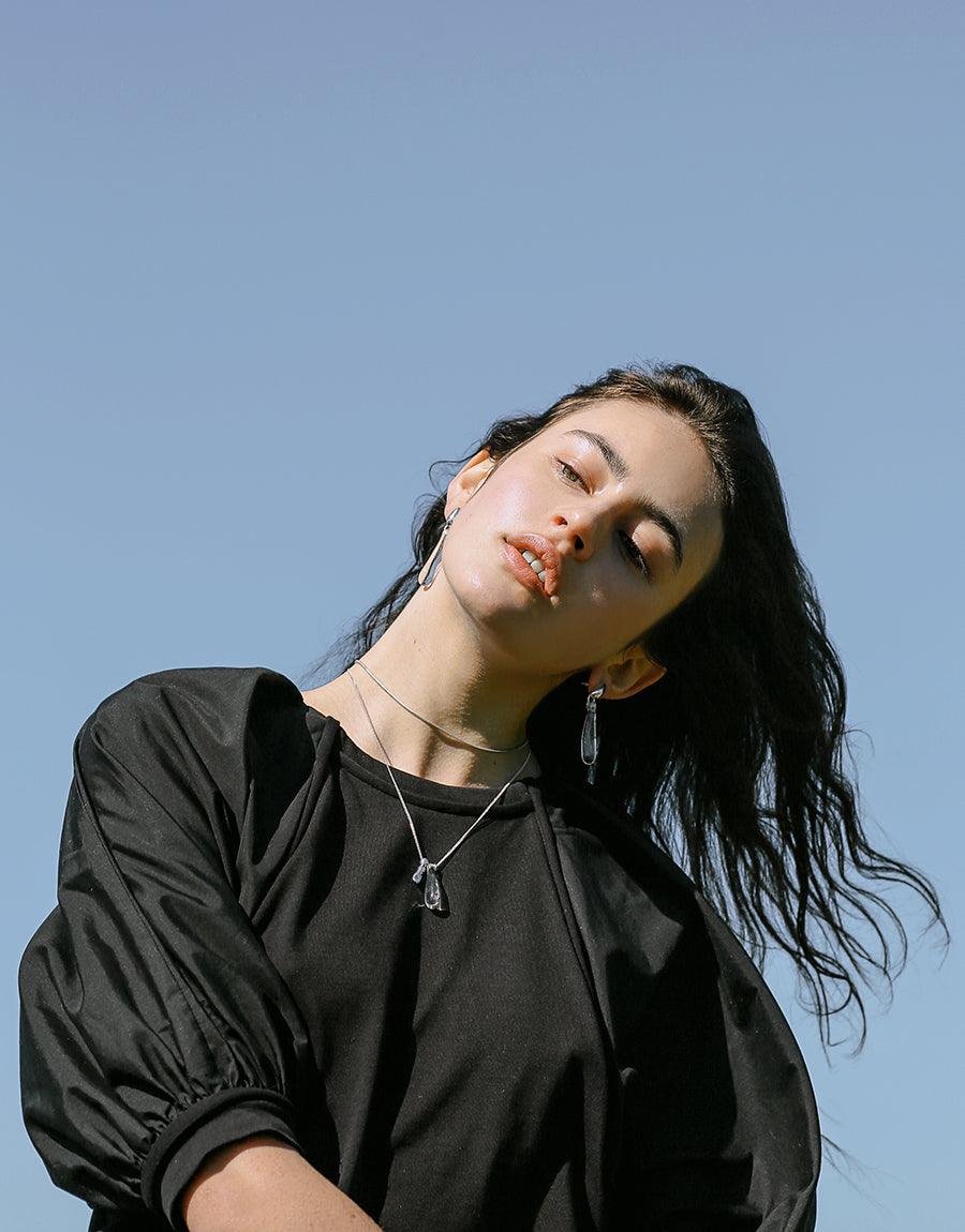 Elegant woman wearing black top and Drop Counts Necklace, featuring glass water drop pendant and wavy metal design. Sustainable jewelry showcasing eco-friendly craftsmanship against blue sky background. CLED Drop Counts Necklace.