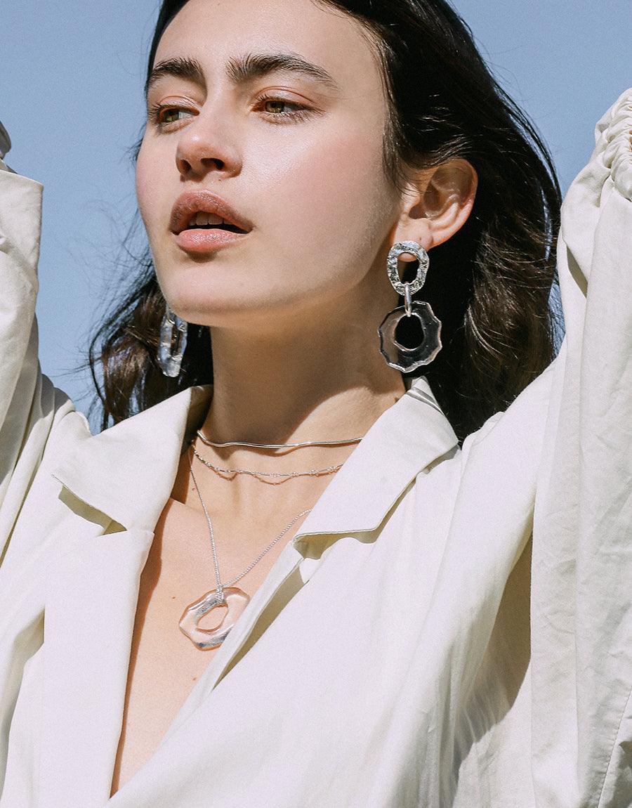 Close-up of model wearing High Tide Necklace, featuring delicate sterling silver chain and organic-shaped eco glass pendant. Paired with white blazer and statement earrings, showcasing versatile ocean-inspired design. Handcrafted sustainable jewelry by CLED.