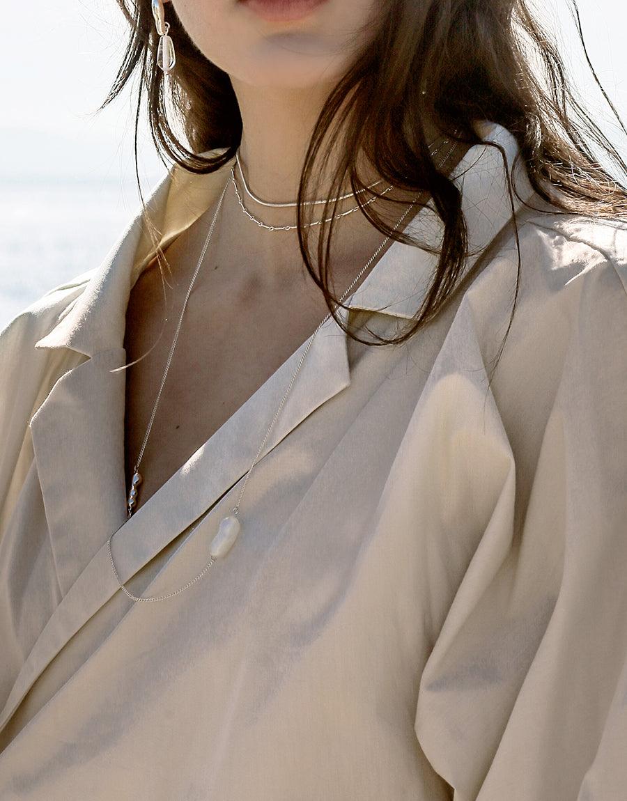 Elegant model wearing Shoreline Long Necklace, a delicate sterling silver chain with sustainable pearl and water drop pendant, paired with a cream-colored blazer against a bright background, showcasing the necklace's versatile style. CLED Shoreline Long Necklace.