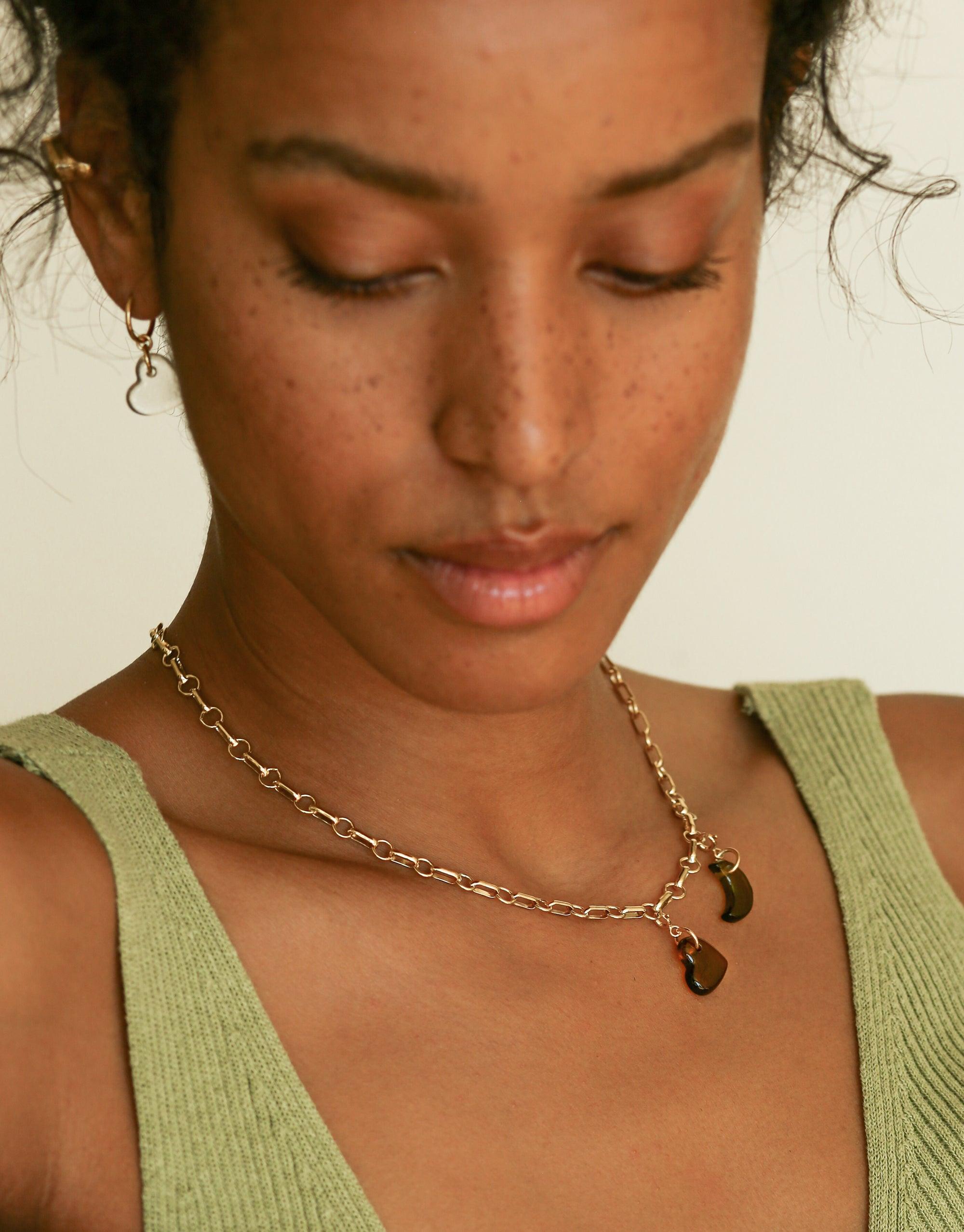 Close-up of elegant gold chain necklace with small pendant charms, worn by model in green top. Versatile and stylish Icon Chain Necklace | B, perfect for customizing with additional charms or wearing alone as a statement piece. CLED