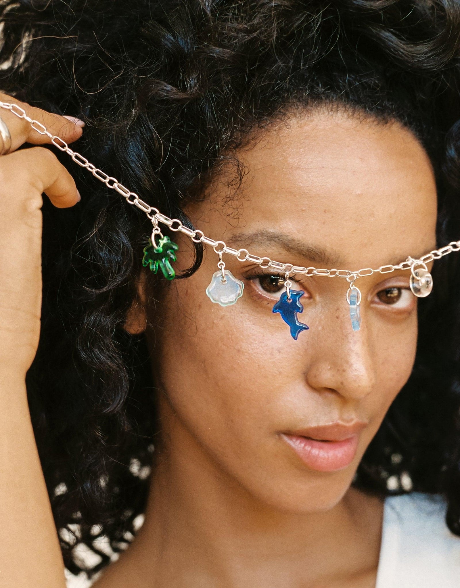 Silver chain necklace with colorful charms including green, blue, and white elements worn by a person with curly dark hair, showcasing the versatility of the Icon Chain Necklace | A. Customizable jewelry for unique style statements by CLED.