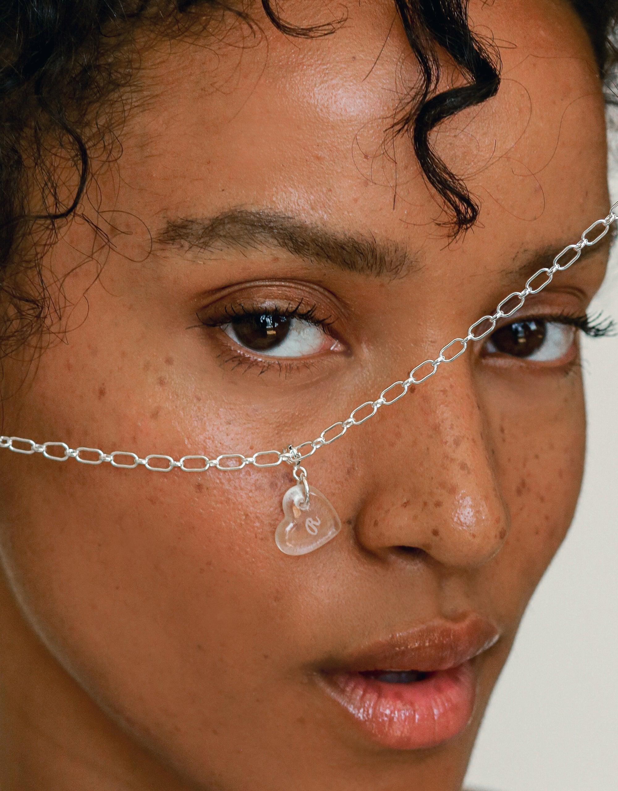Close-up of a delicate silver chain necklace featuring a transparent heart-shaped charm. The necklace rests gently across the bridge of the nose, highlighting the Heart Icon Charm's unique design. Handcrafted jewelry by CLED.