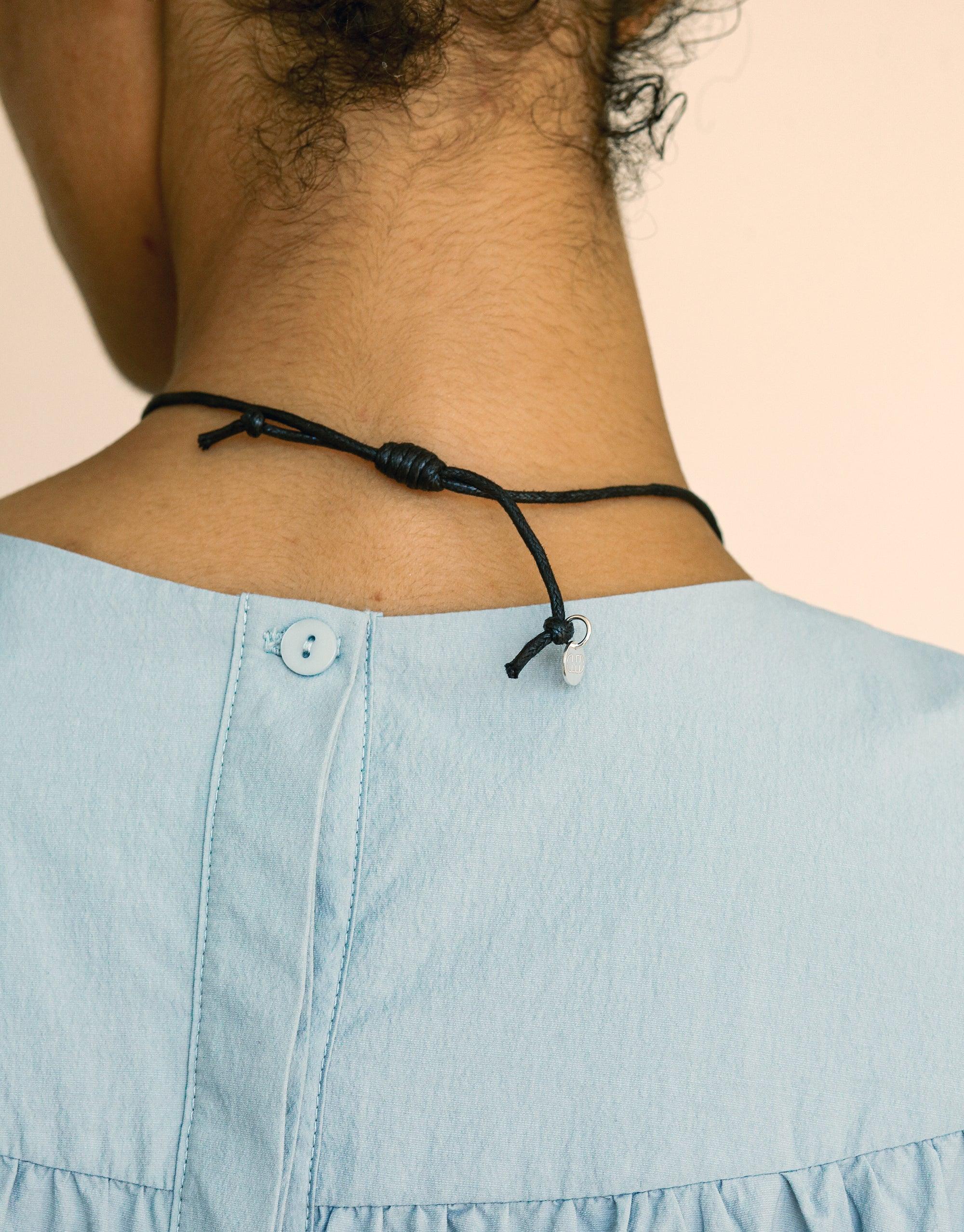 Close-up of a person's neck wearing a black cord necklace with a knot closure, paired with a light blue denim top. The image showcases the Sculptural Large Charm Necklace | C, highlighting its versatile design and complementary style with casual attire. CLED