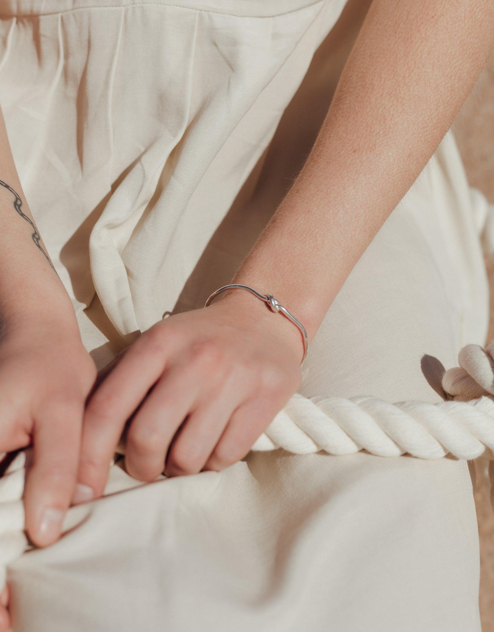 Close-up of hands wearing a sleek silver Single Rope Hook Bracelet against a white fabric backdrop. The bracelet's minimalist design mimics the shape of a twisted rope, showcasing its unique canyon-inspired form. Handcrafted in Los Angeles, this CLED Terrain Collection piece offers an edgy, versatile accessory.
