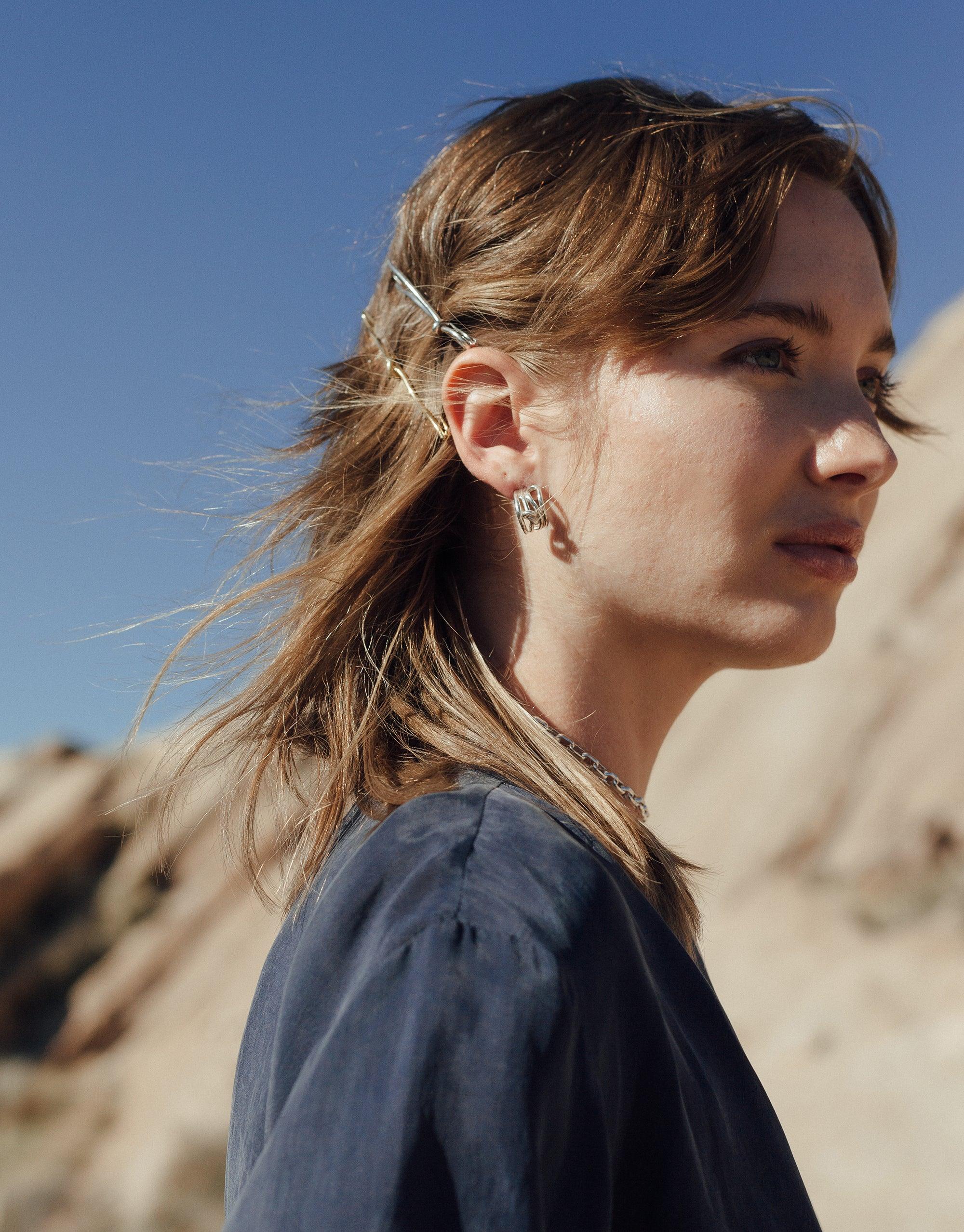 Profile view of a person with windswept hair wearing a dark top outdoors, showcasing a stylish Double Rope Hair Barrette against a desert backdrop. The barrette's elegant design complements the natural terrain, embodying the Terrain Collection's canyon-inspired aesthetic by CLED.