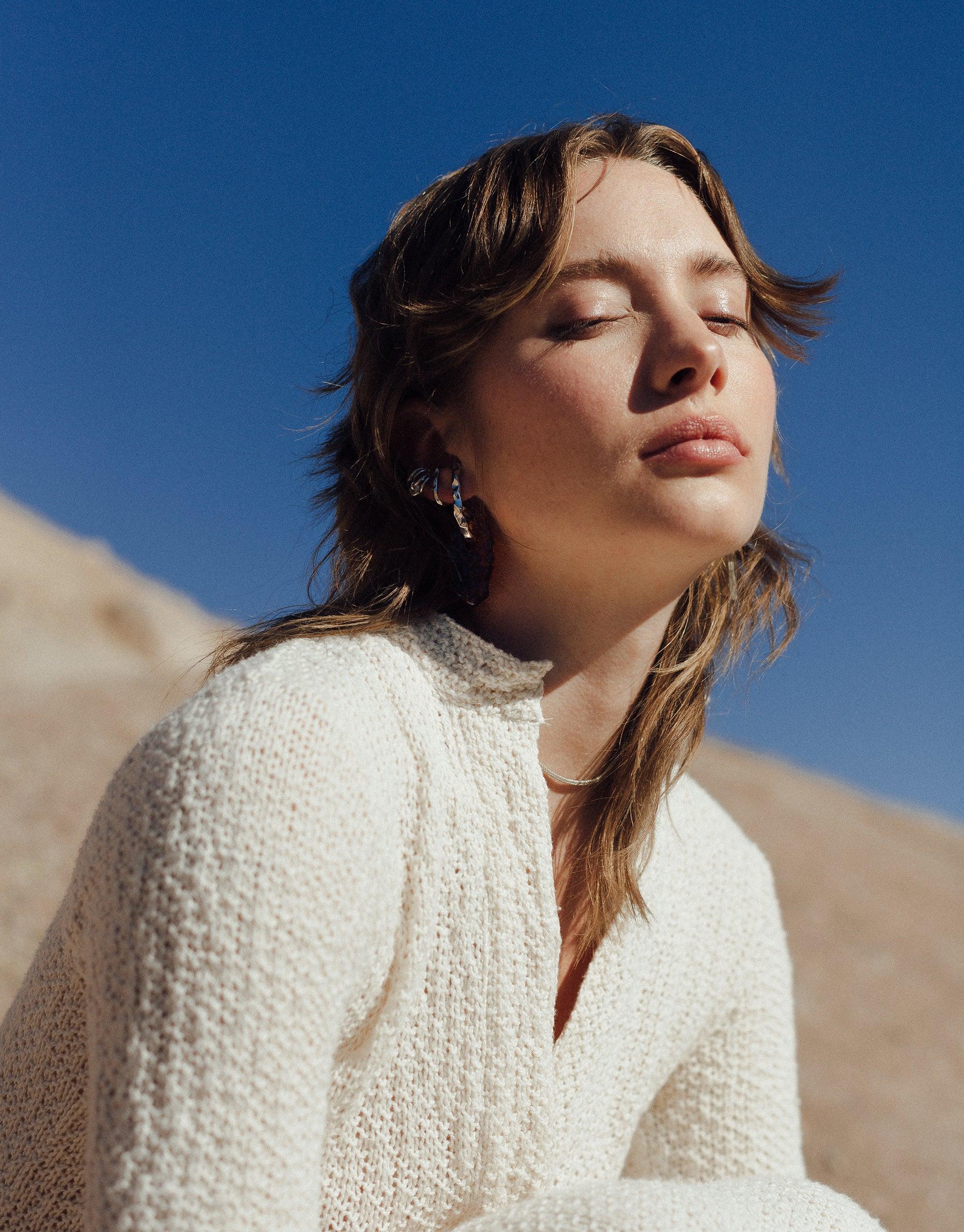 Close-up portrait of a person wearing Rock Spire Earrings, showcasing the unique curved design with glass drops against a backdrop of blue sky and rocky terrain. The earrings complement the natural setting, embodying the Terrain Collection's canyon-inspired aesthetic. CLED Rock Spire Earrings.