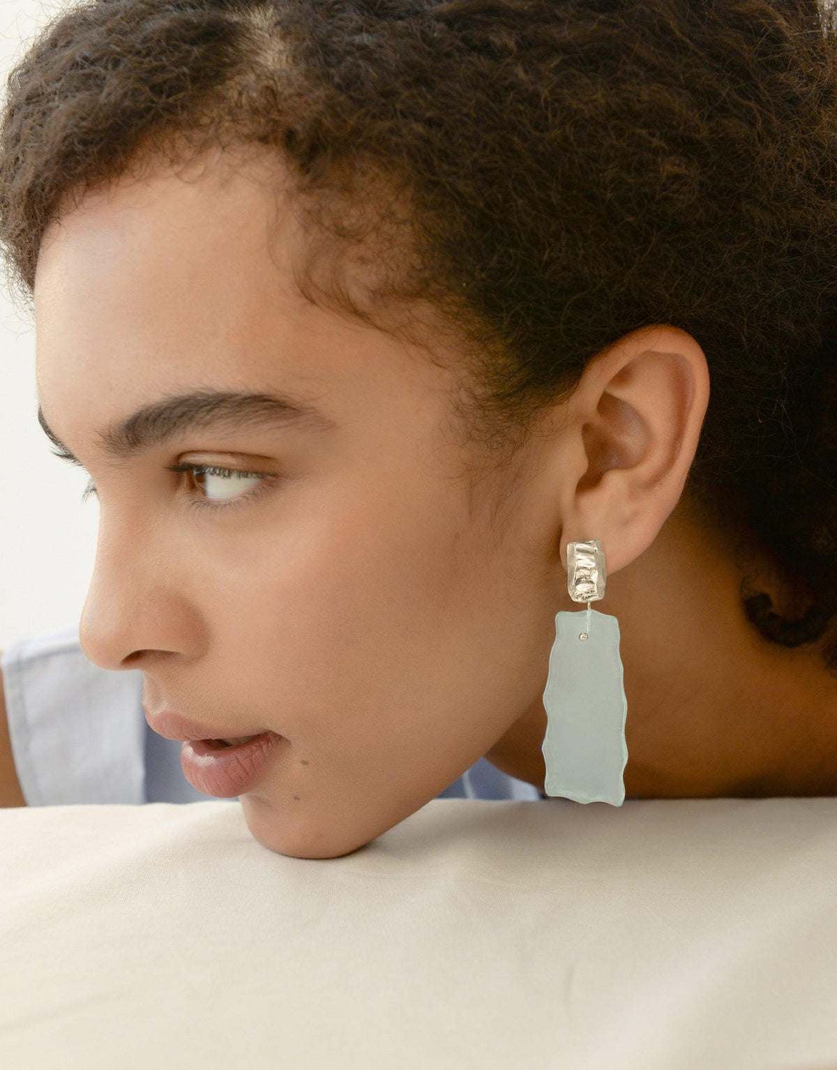 Close-up profile view showcasing Afloat Earrings, featuring a pale blue organic glass pendant suspended from a metallic stud, inspired by melting glaciers. The earring elegantly contrasts against the model's skin, highlighting its unique shape and eco-friendly design. CLED Afloat Earrings.