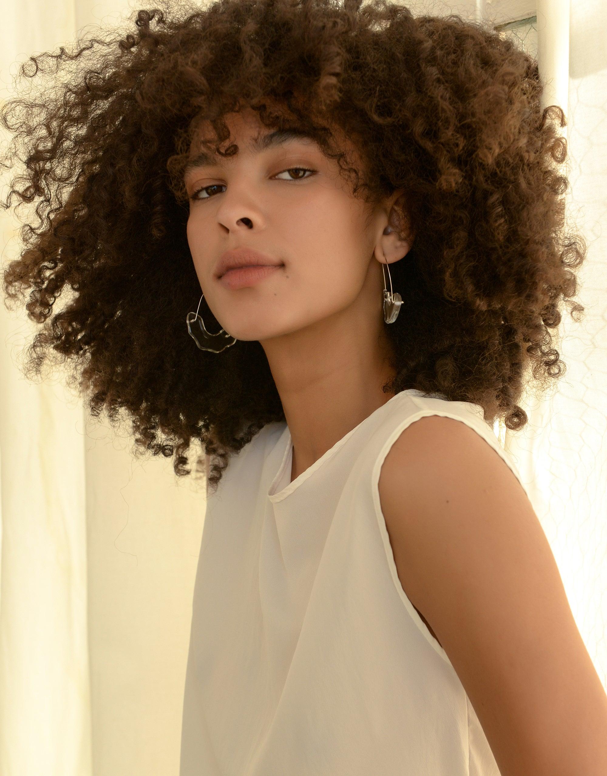 Model with curly hair wearing Polar Arch Hoop Earrings, showcasing elegant design inspired by icebergs. Sustainable jewelry from Floating Glacier Collection, highlighting climate change awareness. Eco-friendly accessories by CLED.
