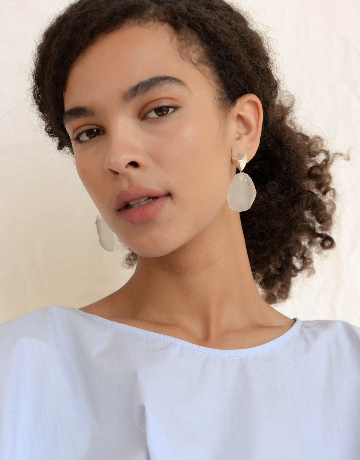 Tip of the Iceberg Earrings: A woman with curly hair showcases sterling silver, matte-finish earrings inspired by icebergs, featuring hand-formed Eco Gems repurposed from discarded glass.