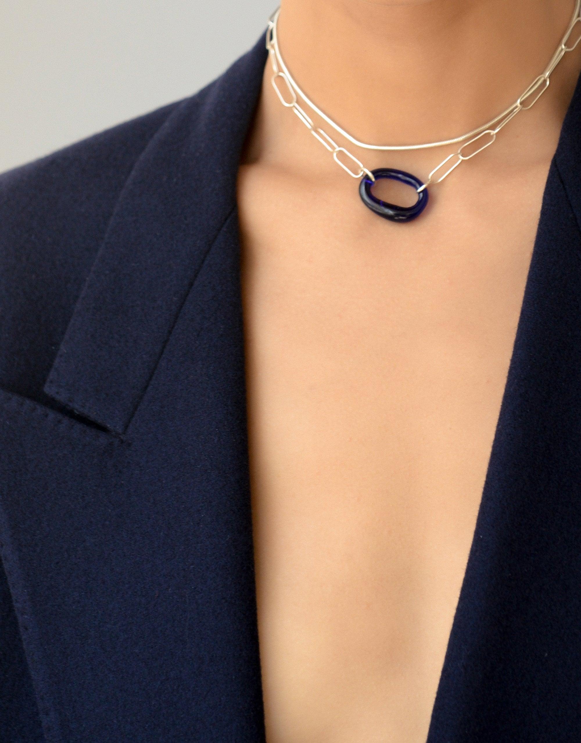 The Day Loop Necklace features an oval chain with a blue Eco Gem pendant, worn by a woman, reflecting CLED's eco-conscious craftsmanship using repurposed glass.